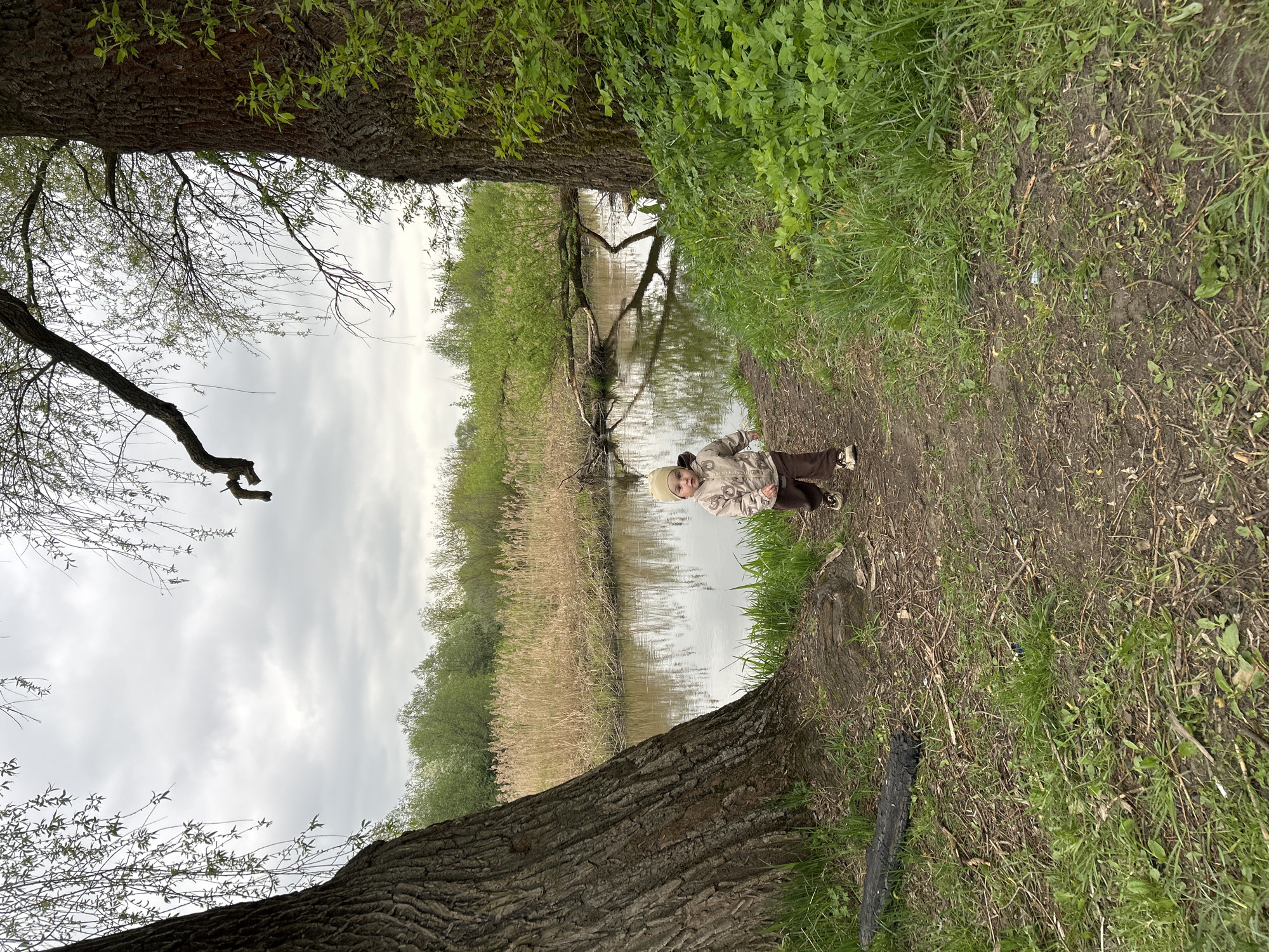 The photograph depicts a child standing on a muddy path alongside a body of water, possibly a river or stream. The child is wearing a light-colored jacket and dark pants, and has a knit cap on their head. The path is bordered by grass and foliage on one side, and the water's edge on the other. Two large trees frame the scene, one on each side. The sky is overcast. The presence of reeds and other aquatic vegetation suggests a natural, possibly marshy environment.
Although no explicit details confirm the location as Klin, Russia, the presence of trees, bodies of water and marshy areas in the image are compatible with the general landscape of that area. More specific identifiers (e.g. characteristic architecture, signage) would be needed to provide positive identification of the location.