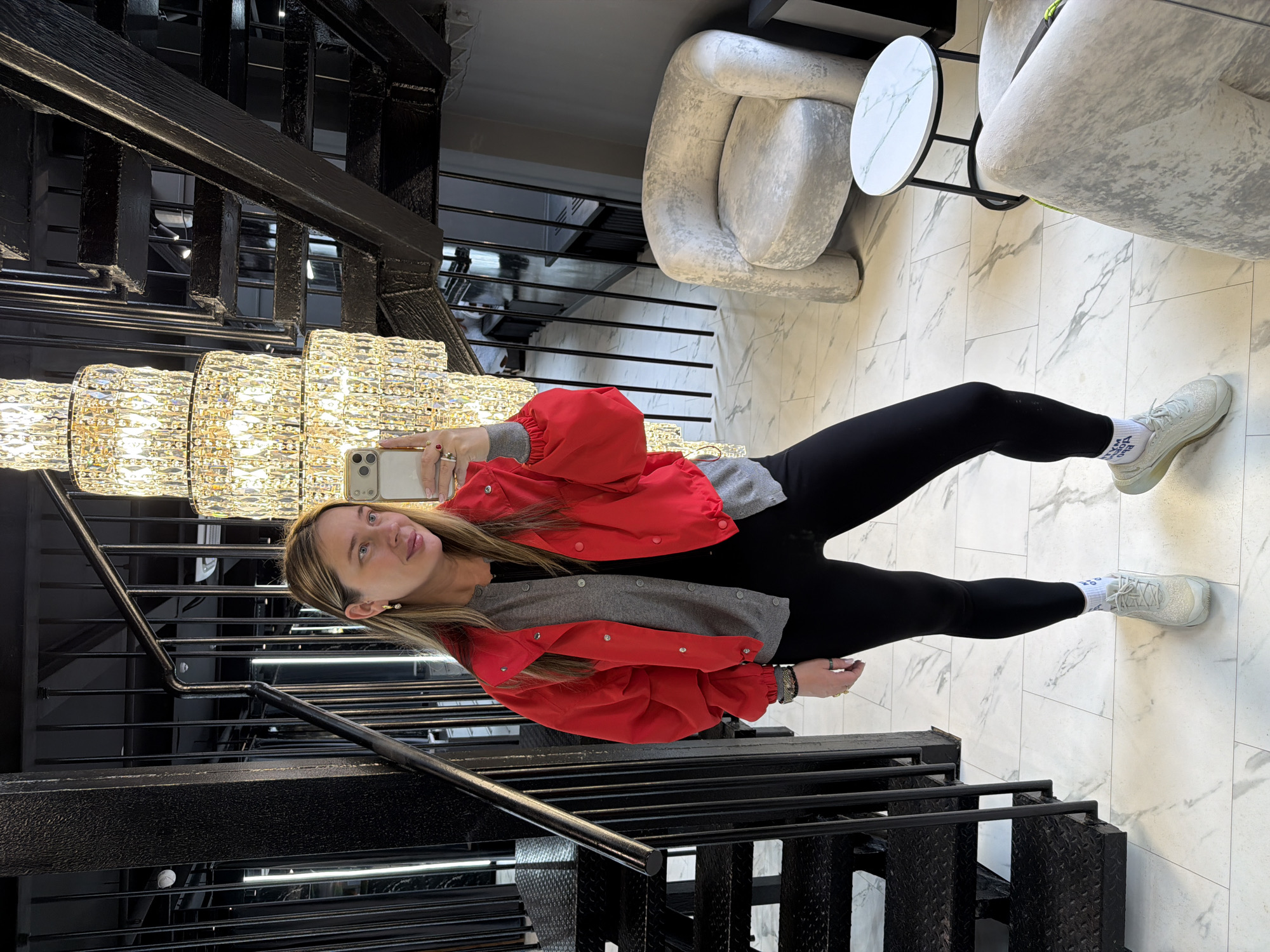 A woman is taking a selfie in an upscale indoor location in Moscow, Russia. She is wearing a red jacket, gray sweater, and black leggings with light-colored sneakers. Behind her is a large, multi-tiered crystal chandelier and a black metal staircase. In the background are modern, light-colored chairs and a small round table with a marble top, suggesting a high-end interior design. The floor is covered in large white tiles with gray veining. The scene suggests an upscale commercial or residential space.