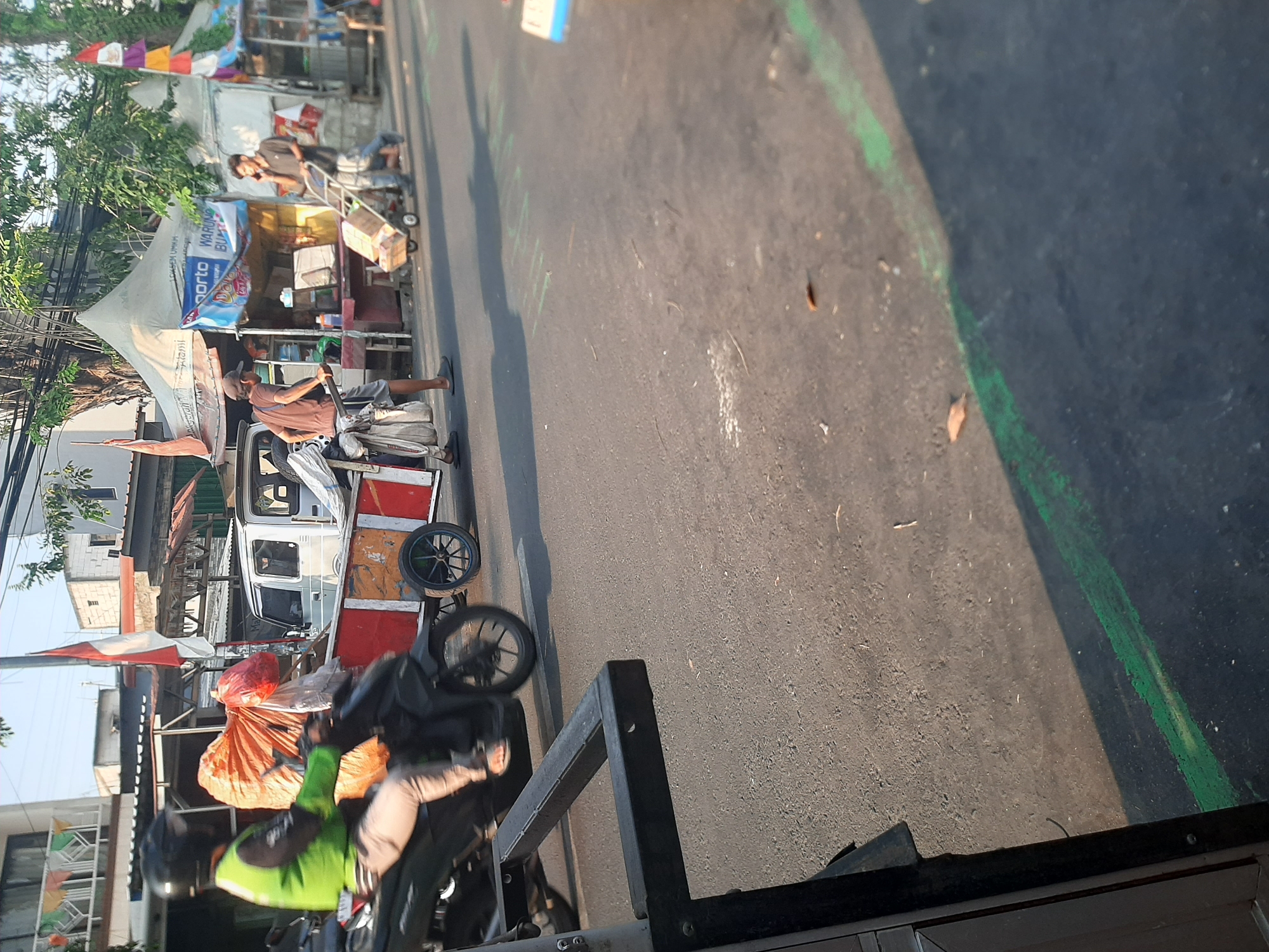 The image depicts a street scene in Central Jakarta, Indonesia, during daylight. The foreground is dominated by the asphalt road, with a portion of a dark-colored railing or barrier on the left. A person wearing a green vest and a helmet is seated on a black motorcycle, which is pulling a trailer. The trailer is partially visible, appearing to be red and yellow with various items, including large orange and white bags, loaded onto it.
In the middle ground, several individuals are present. One person, wearing a brown shirt and a cap, is pushing a cart filled with items, including what appear to be tires and bags. Another person, wearing a dark shirt, is talking on a mobile phone while pushing a wheeled cart laden with cardboard boxes. Behind them, there are several temporary structures, possibly market stalls or food vendors, with awnings and banners. One banner displays "porto WARUNG BUMI." Indonesian flags are visible, one prominently displayed on a pole. The environment suggests a bustling street with commercial activity. The background shows buildings and trees, with clear daylight indicating it is daytime.