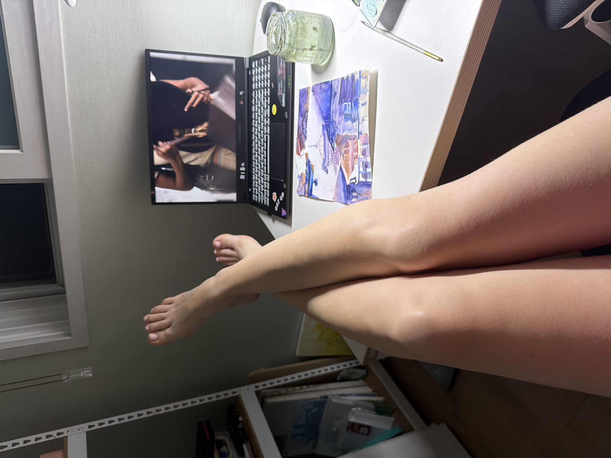 In a room in Dongducheon-si, South Korea, a person's legs are crossed, with their feet in the foreground. A laptop is open on a desk, displaying a video of someone holding items. Beside the laptop is a watercolor painting of buildings and water. A glass jar containing a green liquid and a paintbrush are also on the desk. In the background, a shelf unit holds various art supplies. The lighting suggests it is either early morning or late evening.