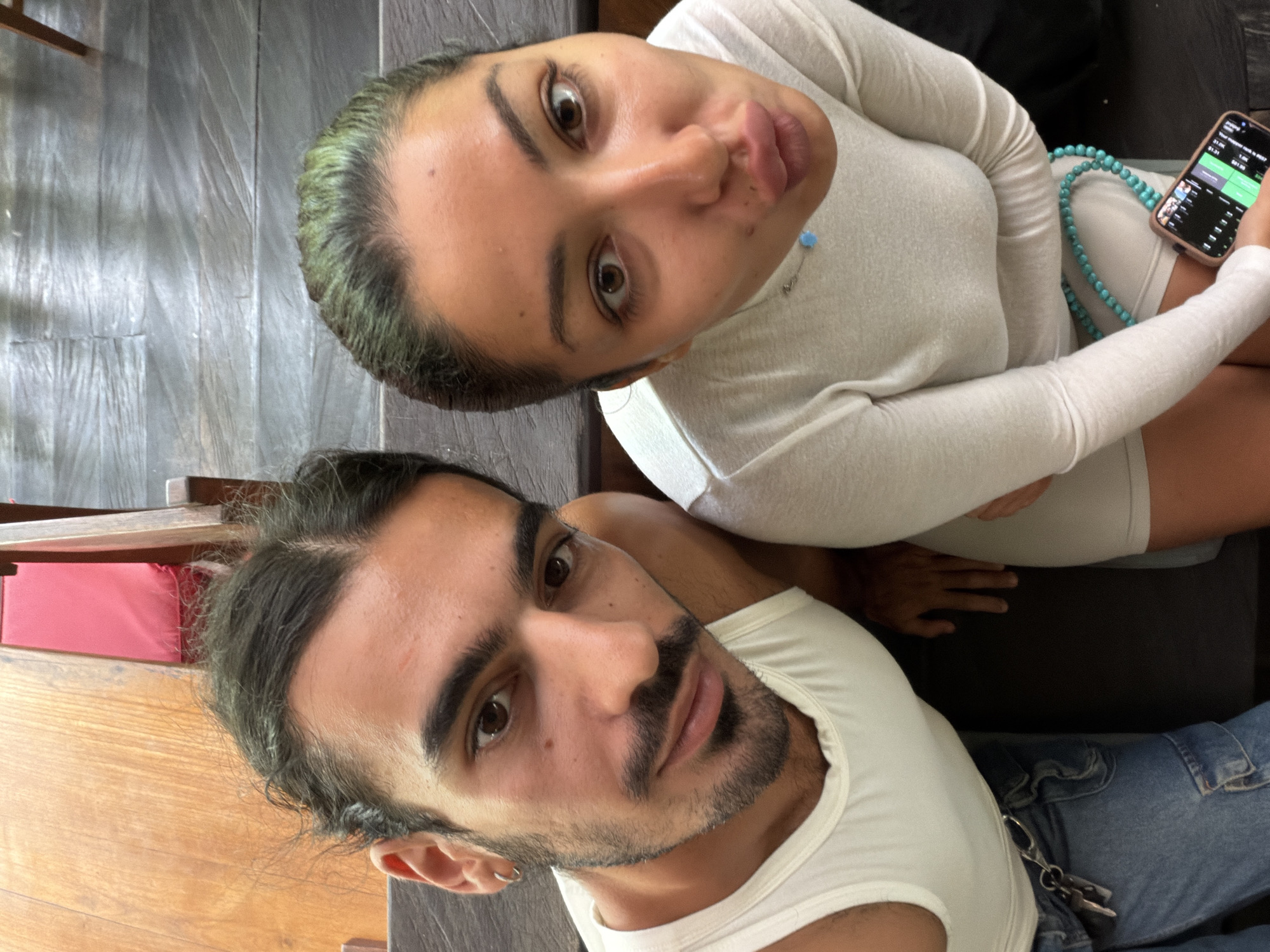 Two people are posing for a selfie taken from a high angle, possibly in North Kuta, Indonesia. A woman with dyed-green hair and brown eyes is pouting into the camera while holding a smartphone. She is wearing a white long-sleeved top and a turquoise beaded necklace. A man with dark hair and a beard is looking at the camera. He is wearing a white tank top and blue jeans. There are wooden and tiled surfaces in the background.