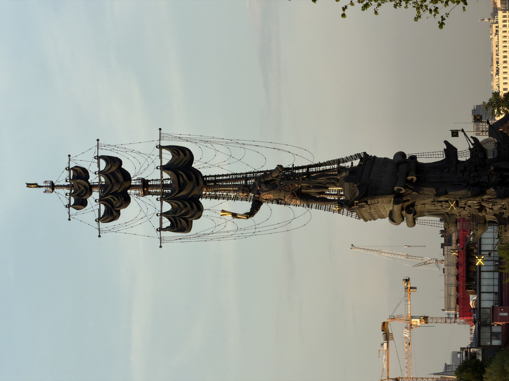 The image shows the Peter the Great Statue in Moscow, Russia. The statue depicts a tall sailing ship with dark sails, rigging, and Peter the Great holding a golden scroll at the bow. Construction cranes and buildings are visible in the background. The sky is overcast. The statue is illuminated, possibly by artificial lights.