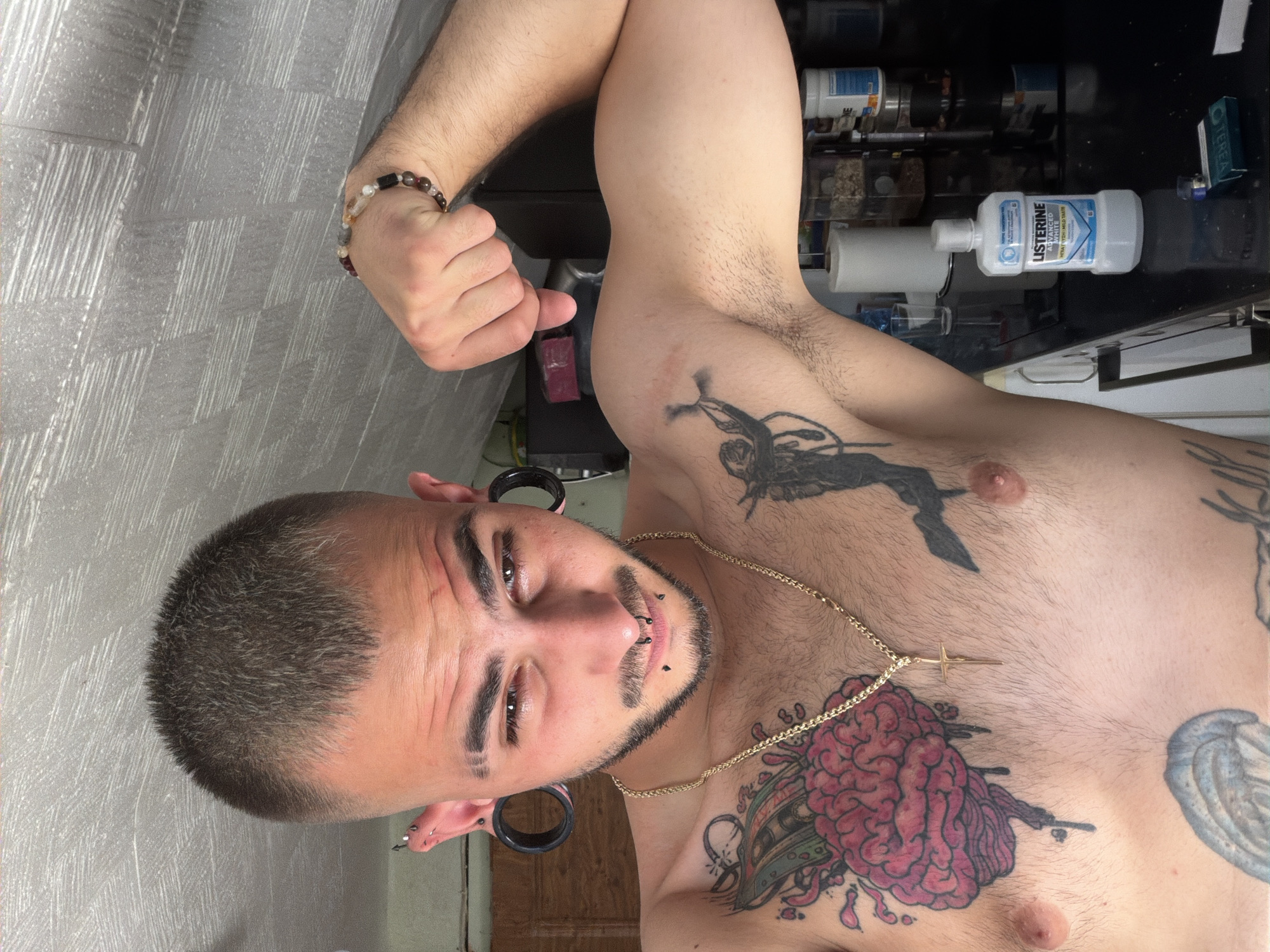 The image displays a shirtless male individual within an indoor domestic setting, identified as being in Plovdiv, Bulgaria. The man, with short, buzzed dark hair, a trimmed beard and mustache, and distinctive shaved lines in his eyebrows, looks towards the camera with a focused gaze.
He is extensively modified with large black ear gauges, multiple ear piercings (some with spike-like jewelry), a septum ring, and two black lip studs. Around his neck hangs a gold chain with a cross pendant, and a beaded bracelet adorns his right wrist. His torso is covered in tattoos, including a large, colorful depiction of a brain with monetary elements on his upper left chest, and a black stylized figure in motion on his upper right arm. Other tattoos are partially visible on his lower body. He is captured posing, flexing his left bicep.
The background features a black countertop holding a bottle of Listerine Advanced mouthwash, a small blue and green packet (resembling heat-not-burn tobacco sticks), and a metallic object. Behind these are a roll of paper towels on a holder, clear containers with dry goods, and various white bottles on higher shelving units. A dark wooden door or cabinet is visible on the left side of the frame. The walls and ceiling are a light grey with a textured finish.