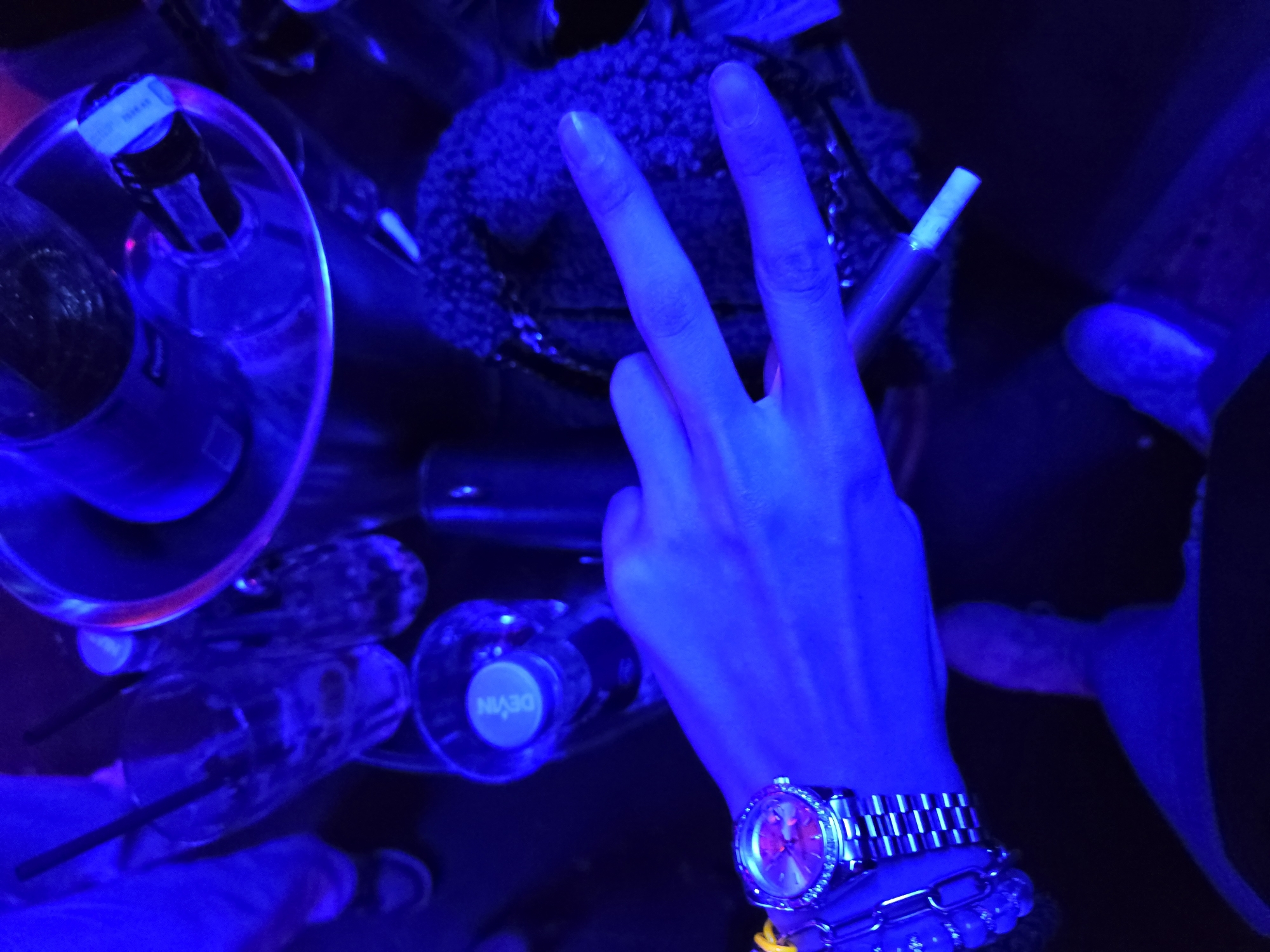 Under blue lighting, the image shows a hand making a peace sign gesture. The hand wears a metal watch and bracelets. Around the hand are bottles of drinks and other objects. The photo does not indicate any specific information that would place it in Varna, Bulgaria.