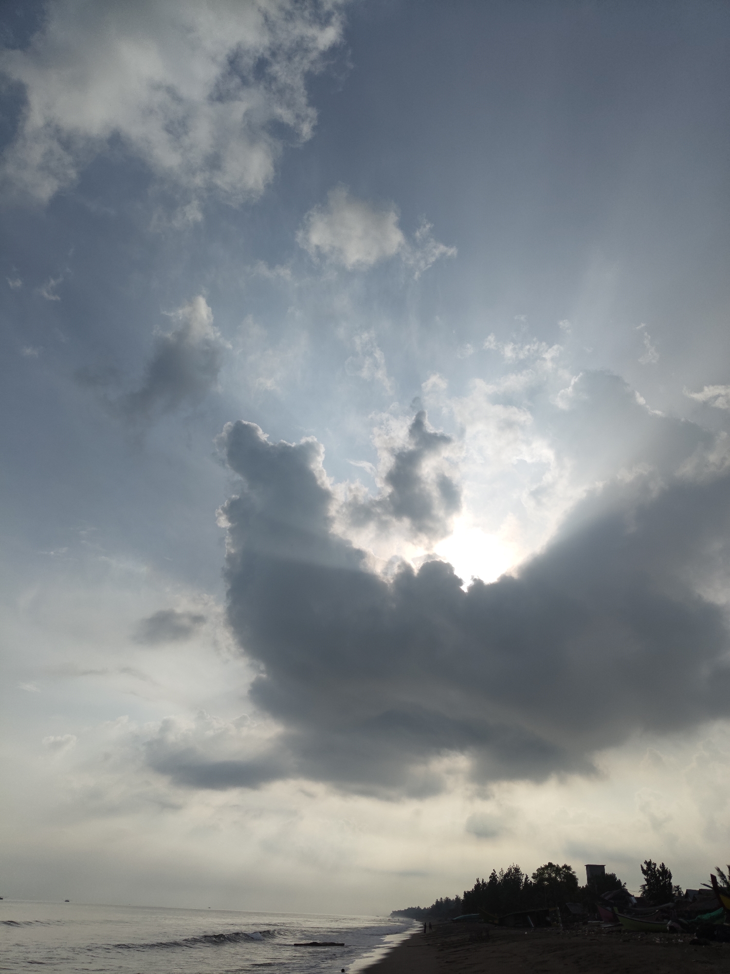 This is a photograph taken at Curehtunong, Indonesia. The image depicts a beach scene with a dramatic sky. The sun is partially obscured by dark, heavy clouds, allowing rays of light to beam through. The ocean is visible in the lower left portion of the frame, with gentle waves reaching the shore. The beach itself is sandy, with some vegetation and a few structures visible in the distance along the tree line. There are no people or animals clearly identifiable in the image, nor are there any apparent actions or interactions taking place. The overall mood is one of a cloudy day with the sun attempting to break through.