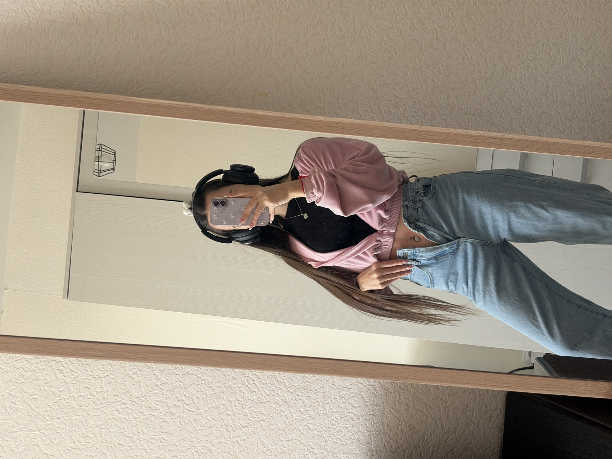 An individual is depicted taking a mirror selfie in an indoor residential setting. The precise location within Saint Petersburg, Russia, is not discernible from the image.
**People:** A single person, appearing female, with long dark hair, stands reflected in a full-length, light wood-framed mirror. Her face is obscured by a smartphone held in her left hand (viewer's right). She wears large black over-ear headphones, a silver necklace with a small pendant, and a red string or bracelet on her right wrist. Her midriff is exposed, revealing a silver navel piercing. Her fingernails are manicured with a light polish.
**Clothing:** The individual is dressed in a black top, a pink cropped zippered jacket with the zipper partially open, and light blue wide-legged denim jeans. The jeans' fly is unzipped and the top button is unfastened.
**Actions:** The person is actively holding a light purple-cased smartphone to capture her reflection in the mirror. Her right hand (viewer's left) rests on the waistband of her unfastened jeans.
**Environment:** The background visible both within and around the mirror consists of a light-colored, textured wall. Reflected in the mirror behind the person are parts of a light-colored wardrobe or closet with two doors, a minimalist wire-frame ceiling light fixture, and a section of a light-colored, multi-drawer cabinet visible to the lower left.