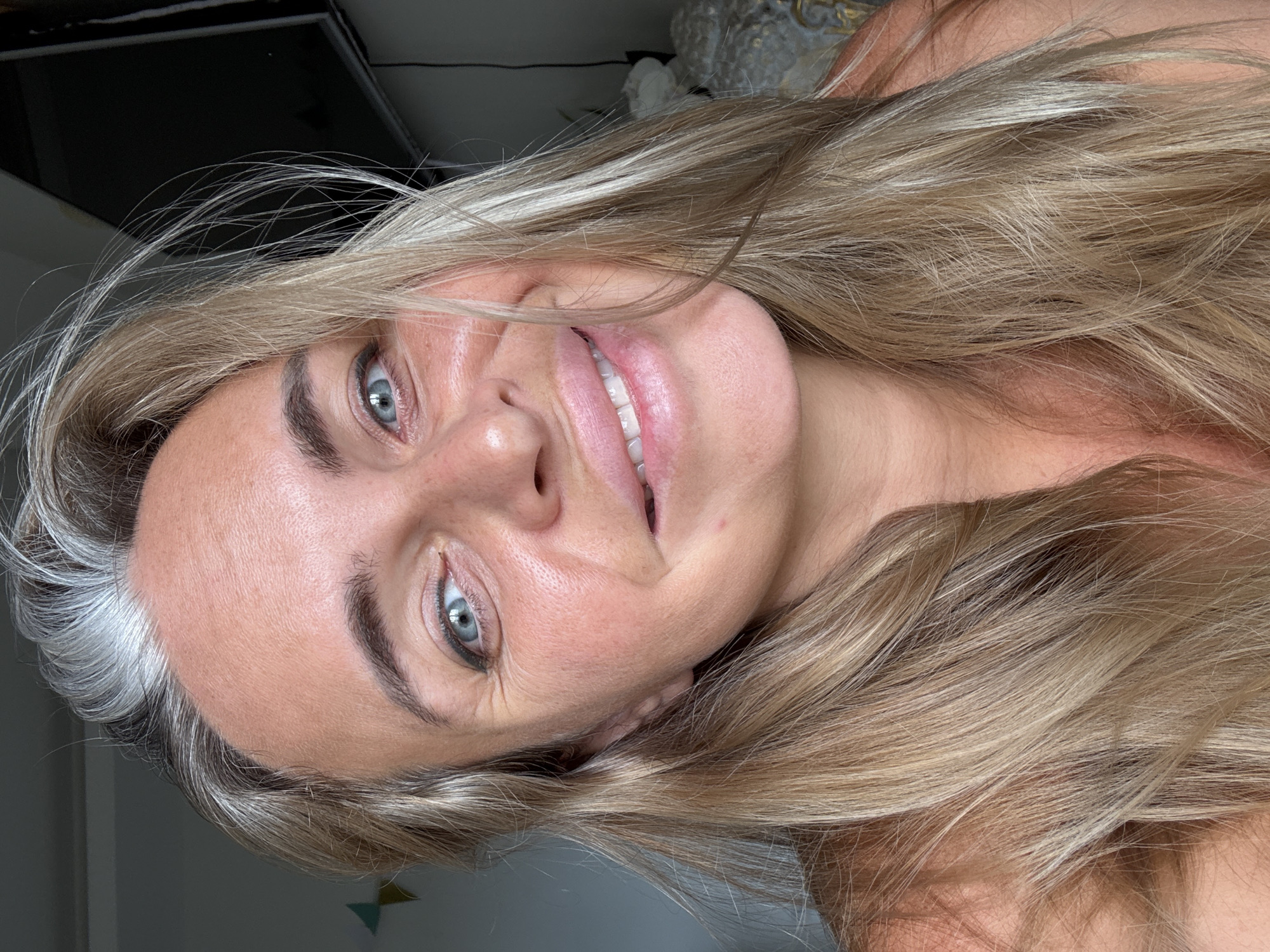 A close-up selfie shows a fair-skinned woman with blonde and grey hair. She is smiling broadly, revealing her teeth. Her eyes are blue-green and her makeup includes eyeliner and lip gloss. The background is blurred, but appears to be indoors with a dark object, possibly a television, on the right and a grey wall on the left. There are no discernible elements that place the image in Ko Samui District, Thailand.