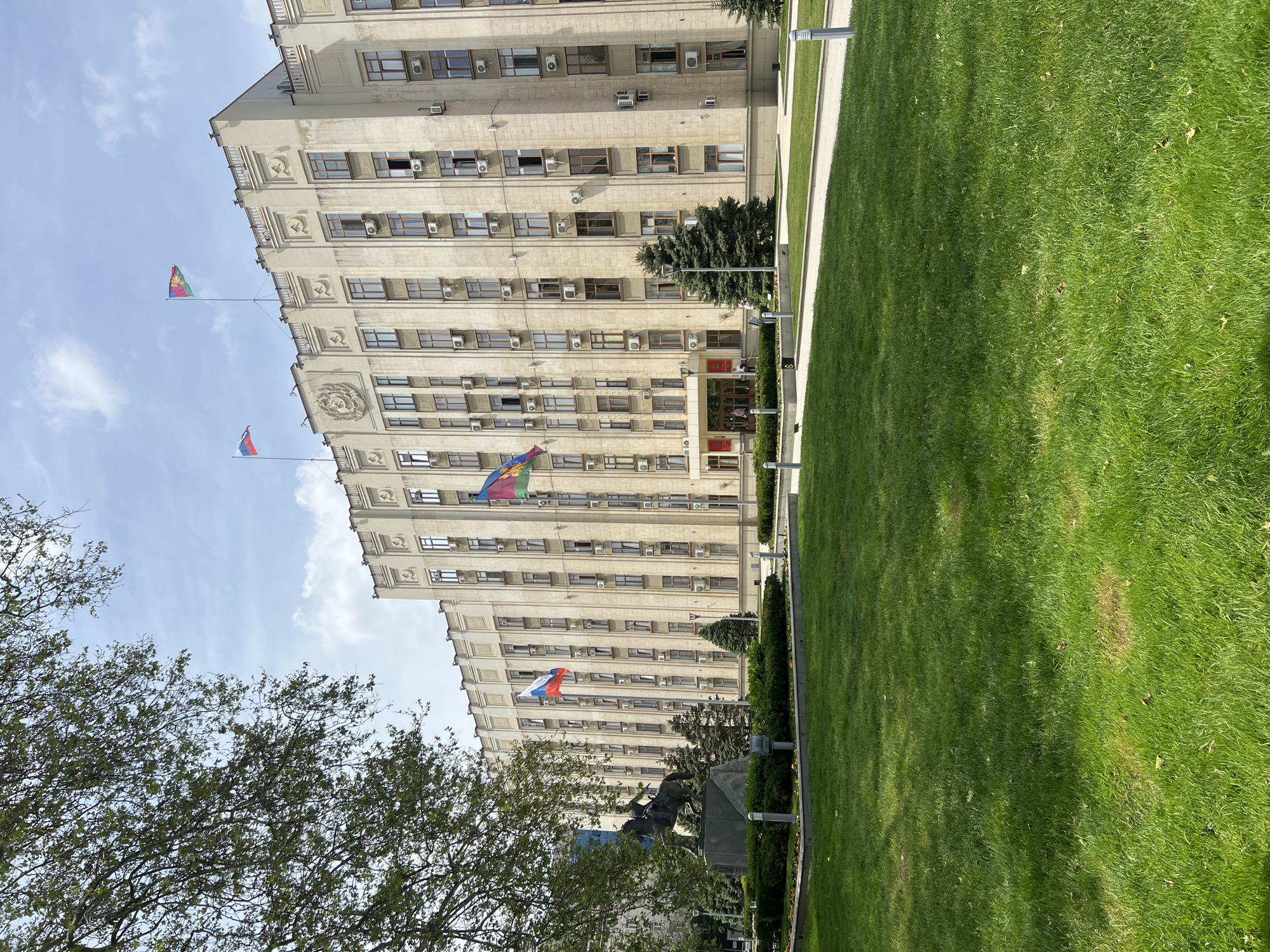 The image depicts the Krasnodar Regional Administration Building in Krasnodar, Russia. The imposing, multi-story, light-colored building stands adjacent to a large green lawn. The Russian flag and the Krasnodar Krai flag are prominently displayed on poles attached to the building's facade. In the foreground, a tree with green foliage partially obscures a view of the building and the lawn. A statue is also visible in front of the building. The sky above is pale blue with scattered clouds.