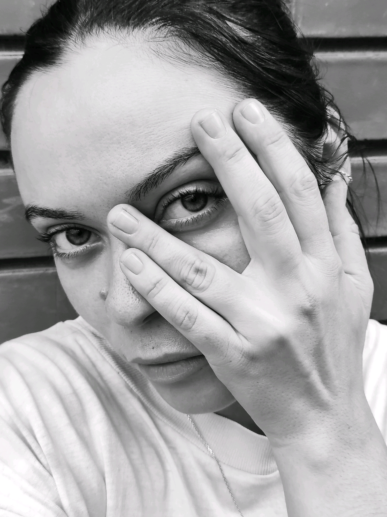 A black and white close-up portrait of a young woman in Фоки, Russia, with her right hand covering the right side of her face, obscuring her eye and nose. Her left eye is visible, looking directly at the camera. She is wearing a white t-shirt. The background is out of focus and appears to be a brick wall. The image is a selfie, likely taken indoors or in an area with artificial lighting, given the evenness of the light on her skin.