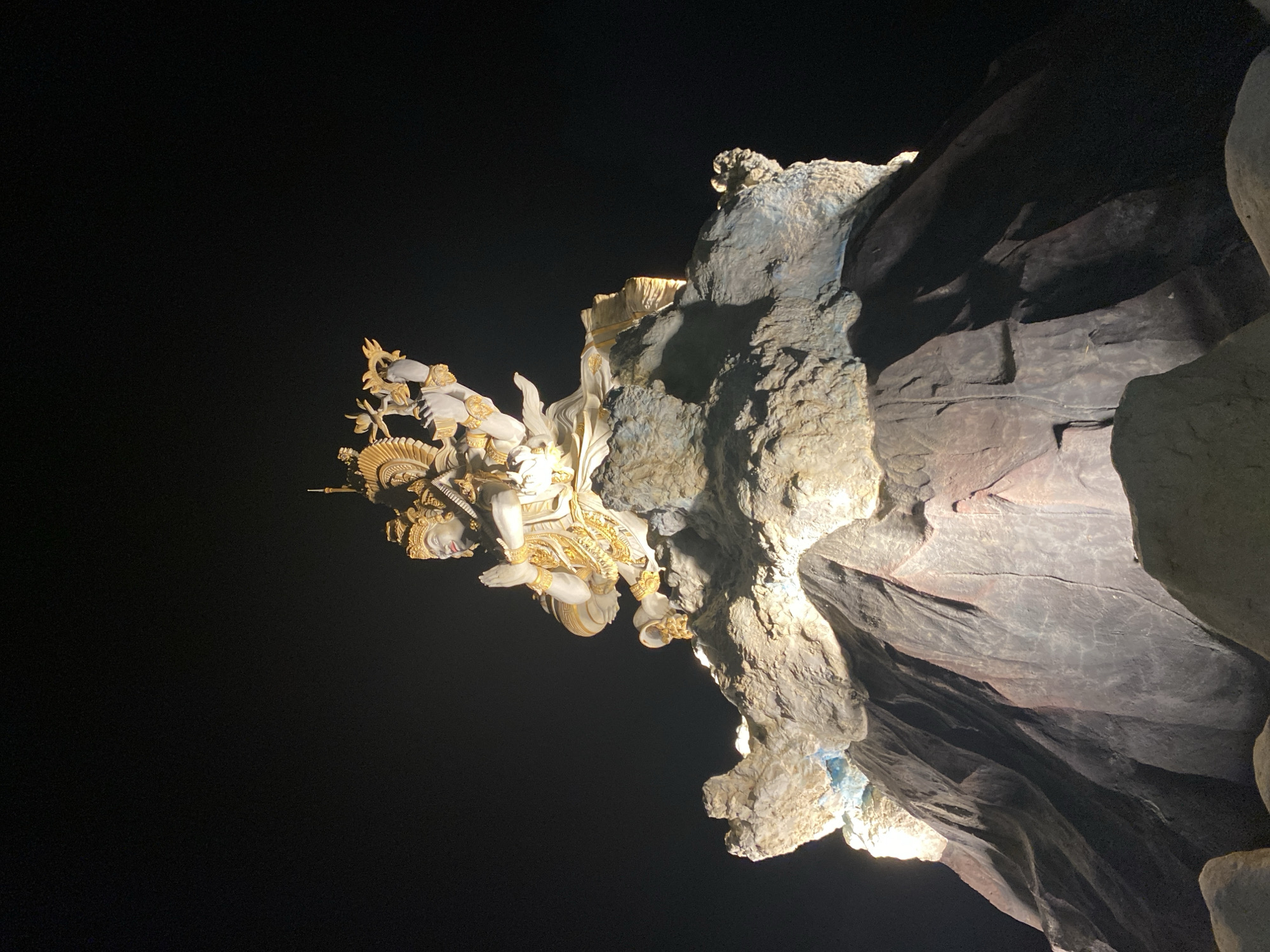 The image depicts a statue of a deity attached to a rocky outcrop against a dark, possibly nighttime, background. The statue is decorated with gold accents and appears to be part of a larger complex within a cave-like structure. The lighting emphasizes the statue and the rock face, creating a contrast against the surrounding darkness. This location is likely in North Kuta, Indonesia, where there are many temples and natural caves.