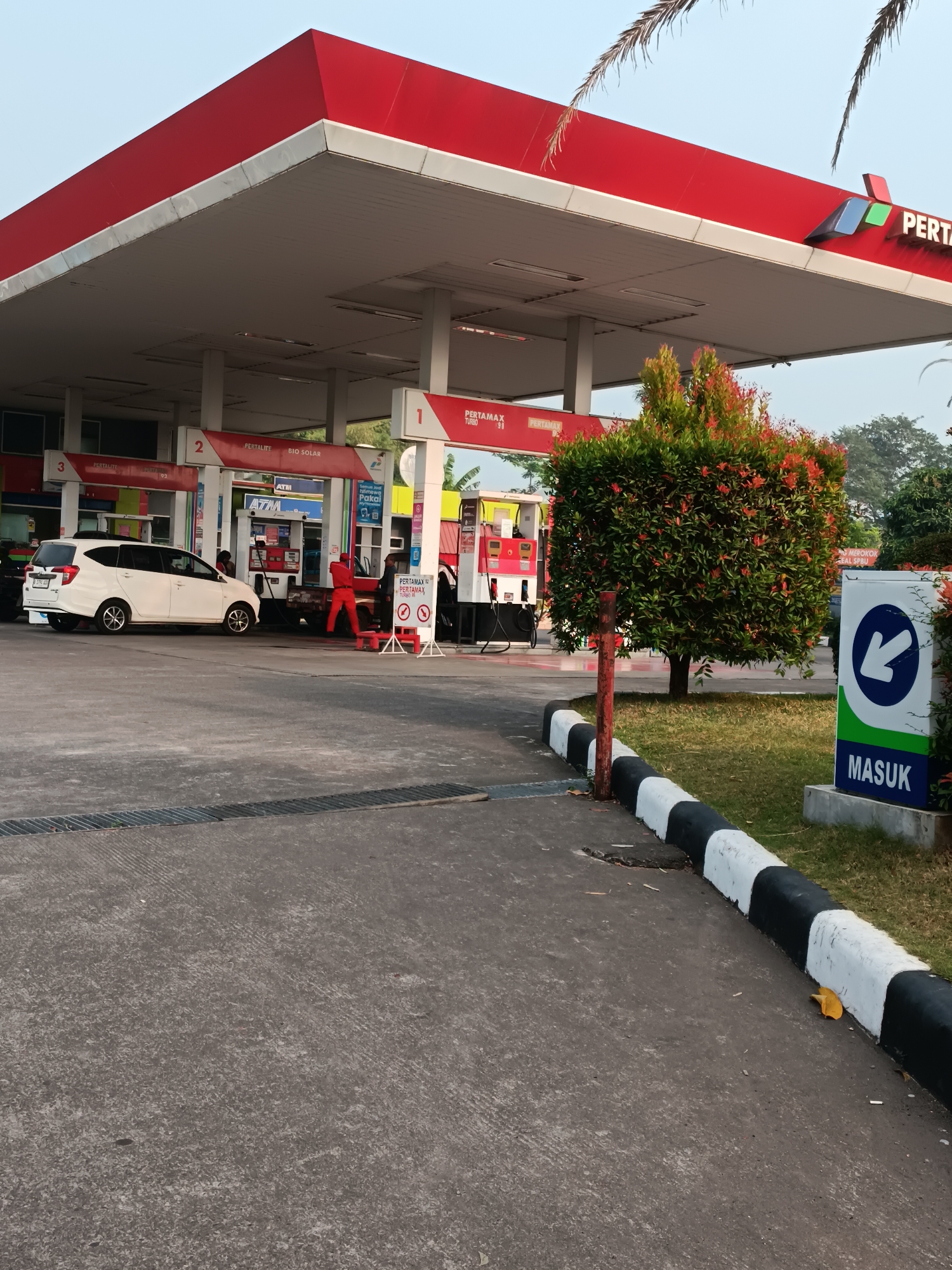 The image depicts a gas station in Pleret, Indonesia, with a bright red roof canopy supported by white pillars. Several fuel pumps are visible, labeled with names like "PERTAMAX TURBO" and "PERTALITE." A white car is parked at a pump, and two individuals, one in an orange uniform and another in dark clothing, appear to be interacting near the pumps. A vibrant green bush with red foliage is situated to the right of the pumps. A sign with an arrow pointing left and the word "MASUK" (meaning "enter" in Indonesian) is also present. The ground is paved with concrete, and a black and white striped curb runs along the edge of the paved area. The sky is clear and appears to be early morning or late afternoon.
