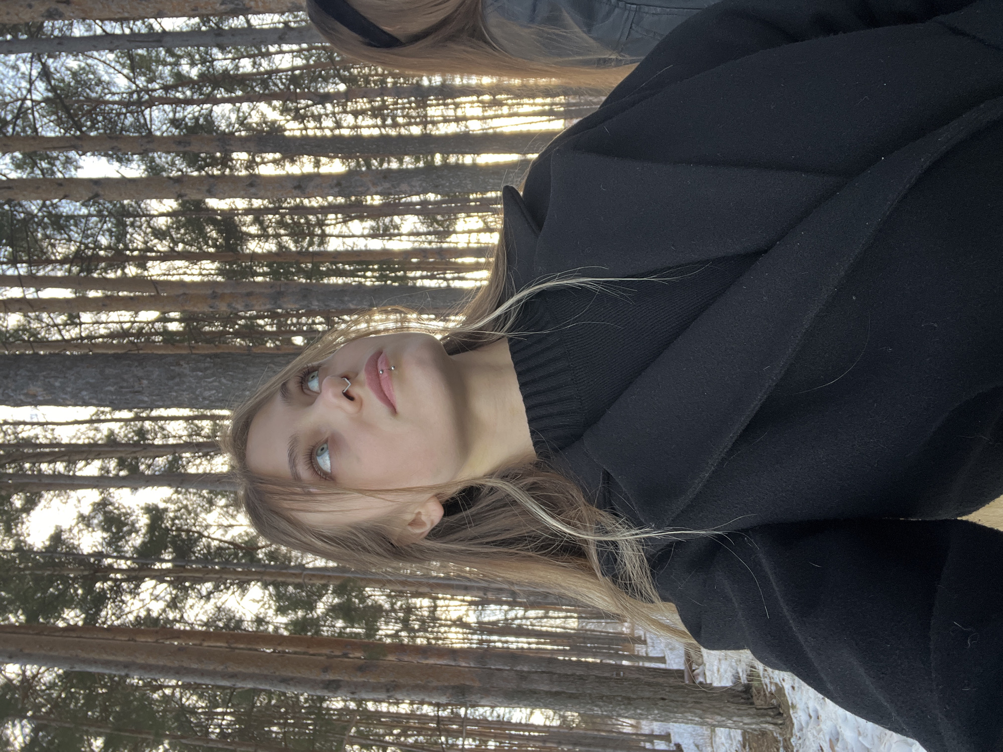The image depicts two individuals in a snowy coniferous forest, located in Chaikovsky, Russia. In the foreground, a young female with long, light-brown hair and light blue-green eyes is captured from the upper chest upwards. She is wearing a black ribbed sweater beneath a black overcoat. Her visible facial features include a silver septum piercing and a silver barbel piercing positioned below her lower lip. Her head is tilted slightly upward and to her left, with her gaze directed into the upper portion of the forest canopy. To the right, partially visible in the midground, is another individual with long brown hair, wearing a black headband and a dark-colored jacket. The forest floor is covered with a thin layer of snow, revealing patches of darker ground beneath. The background consists of numerous tall, slender pine trees, which are heavily backlit by a low sun, creating bright streaks of light between the tree trunks and casting many trunks into silhouette. The sky, visible through the sparse tree canopy, appears bright and hazy.