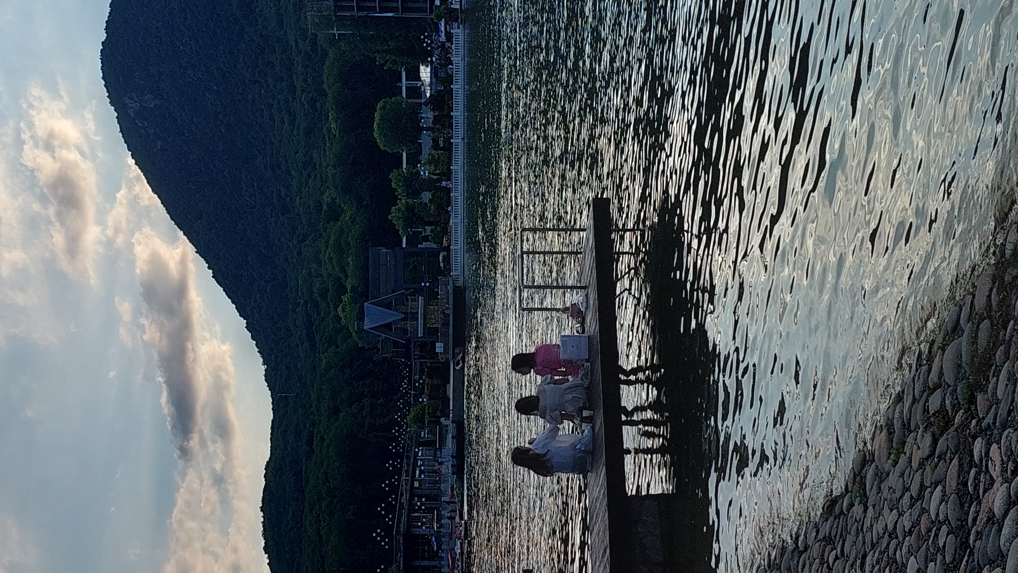 The image depicts a serene waterside scene with a group of people gathered on a wooden dock. The setting is a tranquil lake or river, with a lush green hillside rising dramatically on the left side. The hillside is covered in dense trees and vegetation, creating a sense of natural beauty and seclusion. In the distance, along the shoreline, there are hints of buildings and lights, suggesting a town or resort area.
On the dock, three individuals are seated, seemingly engrossed in an activity. One person, wearing a pink top, is interacting with a laptop, while the other two, in lighter colored clothing, are positioned close by. Their postures suggest a relaxed and comfortable interaction. A metal railing is also visible on the dock, providing a sense of safety and structure.
The water's surface is rippled, reflecting the soft, diffused light of what appears to be late afternoon or early evening. The sky above is a pale blue, with scattered clouds that are tinged with a warm, golden light, indicating either sunrise or sunset. The light on the water creates a shimmering effect, adding to the peaceful ambiance of the scene. The shoreline in the foreground is lined with smooth, rounded stones.
The overall mood conveyed is one of tranquility, relaxation, and connection with nature. The image captures a moment of shared activity and quiet contemplation by the water, with the imposing natural landscape providing a dramatic backdrop.