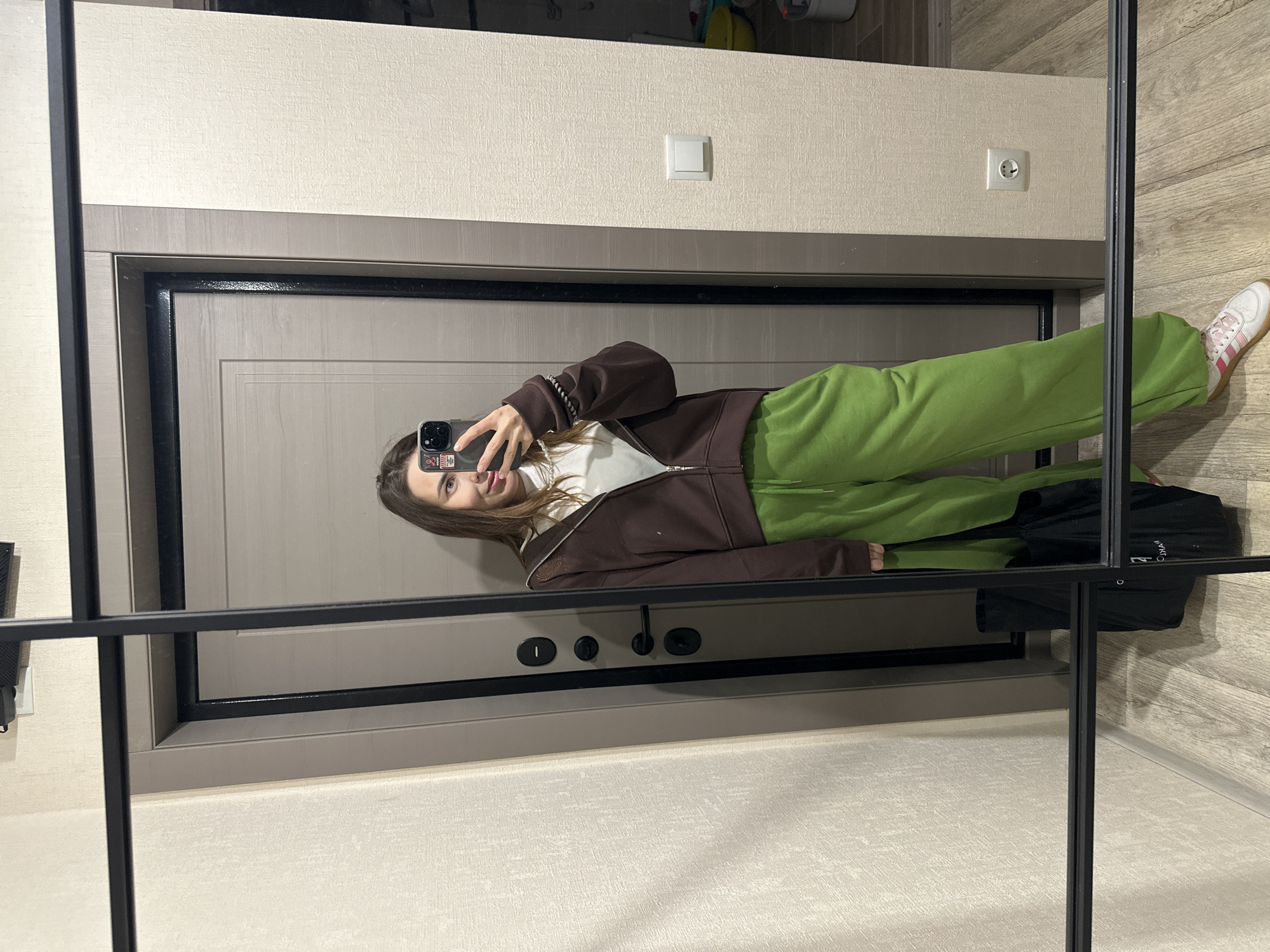 A young woman captures a full-body mirror selfie indoors, holding a dark smartphone to photograph her reflection. She has long brown hair and a subtle, pleasant smile. Her attire consists of a white top, a brown zip-up hoodie or jacket, and baggy bright green trousers, paired with white sneakers featuring pink stripes on the sides.
The setting appears to be a hallway or entryway, reflected in a large mirror with black framing and vertical/horizontal dividing bars. Behind her reflection, a large, modern grey or taupe-colored door with multiple locks and a handle is visible. The room features textured light beige wallpaper on the upper walls, transitioning to a light grey, possibly wood-paneled, section below. The floor is covered in light-colored wood-look planking.
Notable objects include a white light switch and a white electrical wall socket (Type F, common in European countries including Russia), providing a subtle geographical cue. The woman's phone case is black and adorned with a few small stickers, one red with white markings and another white with black designs. On the floor, near her feet, a black bag is partially visible with white text that appears to read "ODYAA" or similar. The time of day is not discernible, but the scene is well-lit.