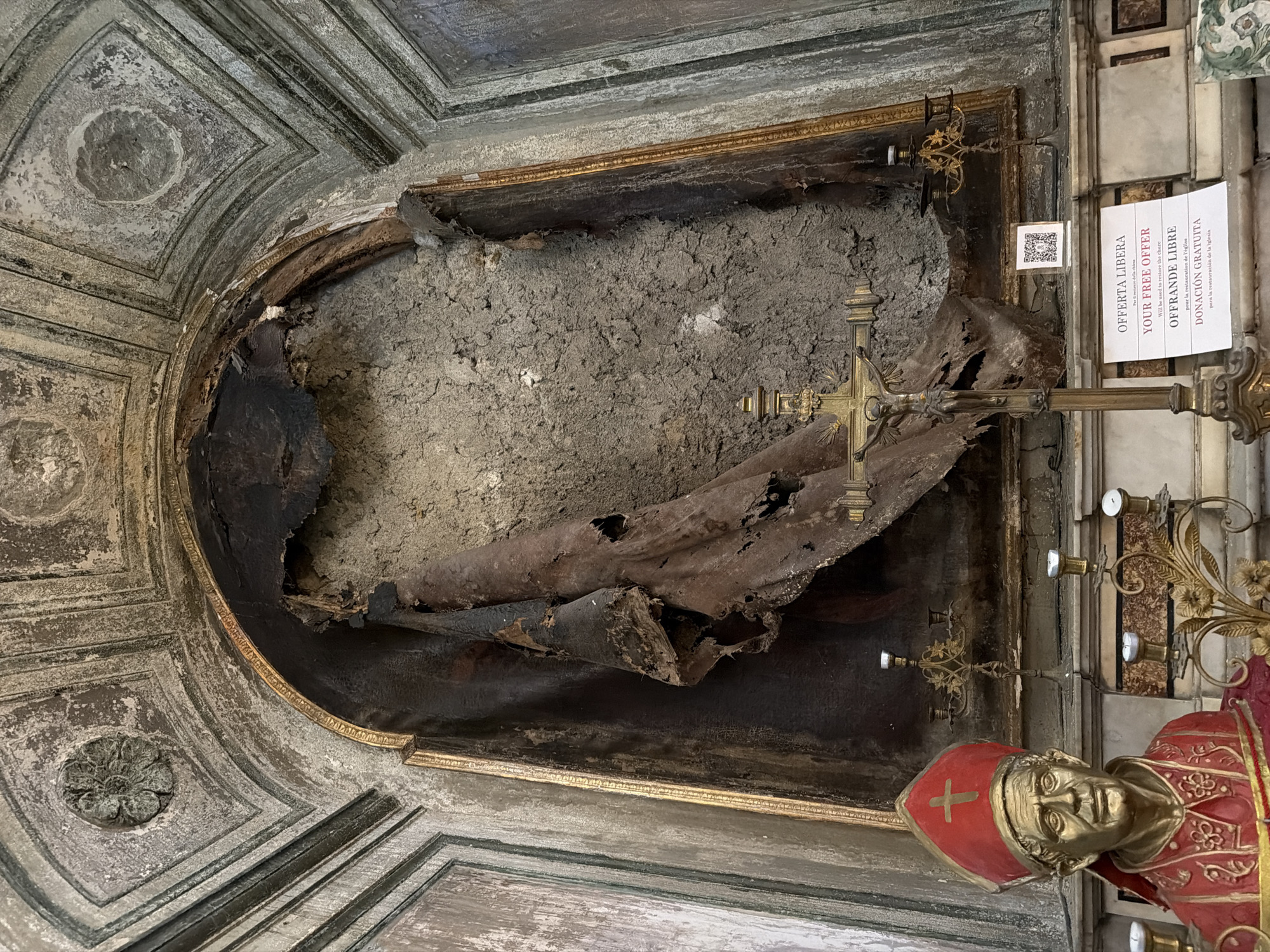The image depicts the interior of a church in Naples, Italy, specifically the Church of San Giorgio Maggiore. The central focus is a damaged fresco within an arched niche. The fresco appears to have suffered significant fire damage, with large portions of the plasterwork and painted surface burned, torn, and hanging in tattered strips. The colors are darkened and muted, with exposed rough plaster visible.
In the foreground, to the left, is a bust of a man dressed in red and gold ecclesiastical robes, wearing a red mitre with a golden cross. This represents Saint Gregory of Nazianzus. In front of the niche, a golden crucifix stands on a pedestal. To the right of the crucifix, a white sign with multilingual text, including "OFFERTA LIBERA" and "YOUR FREE OFFER," is displayed, likely indicating a call for donations for restoration. Several ornate candle holders, some with lit candles, are placed around the lower part of the niche and the foreground. The overall environment suggests a historical religious site that has experienced damage and is undergoing or seeking restoration.