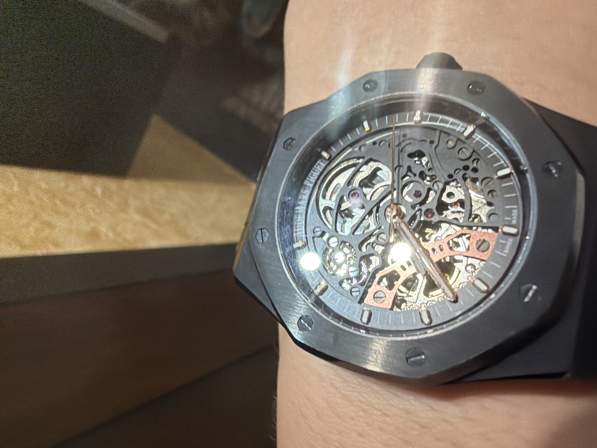 A close-up photograph shows a dark gray Audemars Piguet Royal Oak Openworked watch on a person's wrist. The watch has a skeletonized dial that shows the watch's inner mechanisms. The case has an octagonal shape with visible screws. The watch is positioned in front of a blurred background that appears to be an indoor setting with a combination of dark and light surfaces. The location is not identifiable as Shumen, Bulgaria, from the information contained within the image.