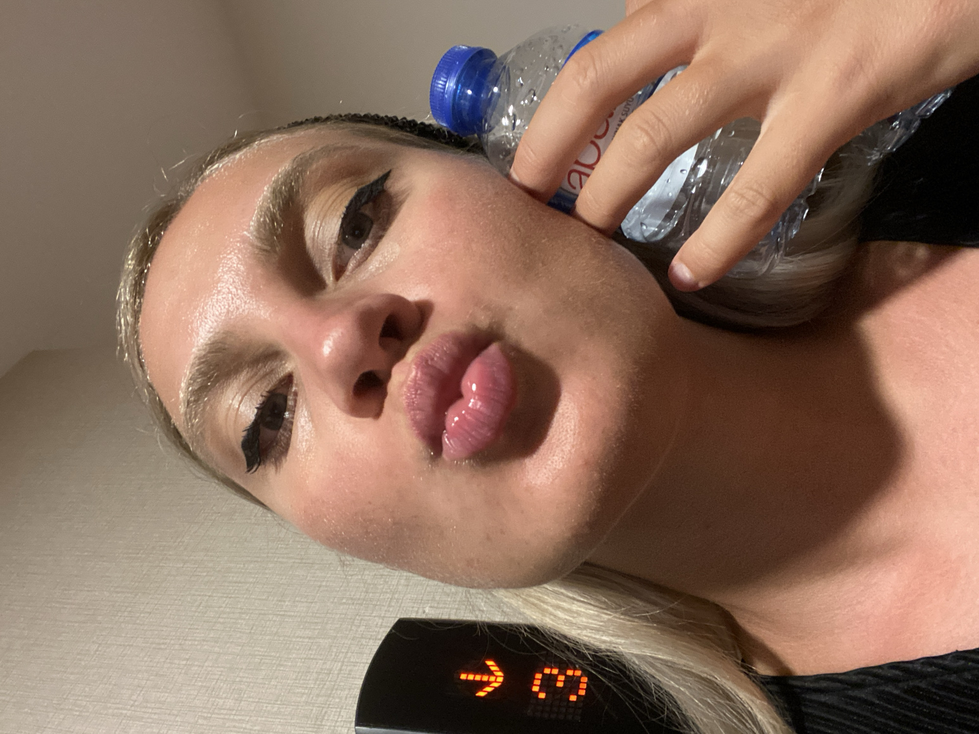 In Aksu, Turkey, a young woman with blonde hair and prominent eyeliner holds a water bottle and poses for a selfie. An elevator panel with an illuminated downward arrow and the number '3' is visible to her left. The scene appears to be an indoor setting, possibly an elevator lobby or hallway.