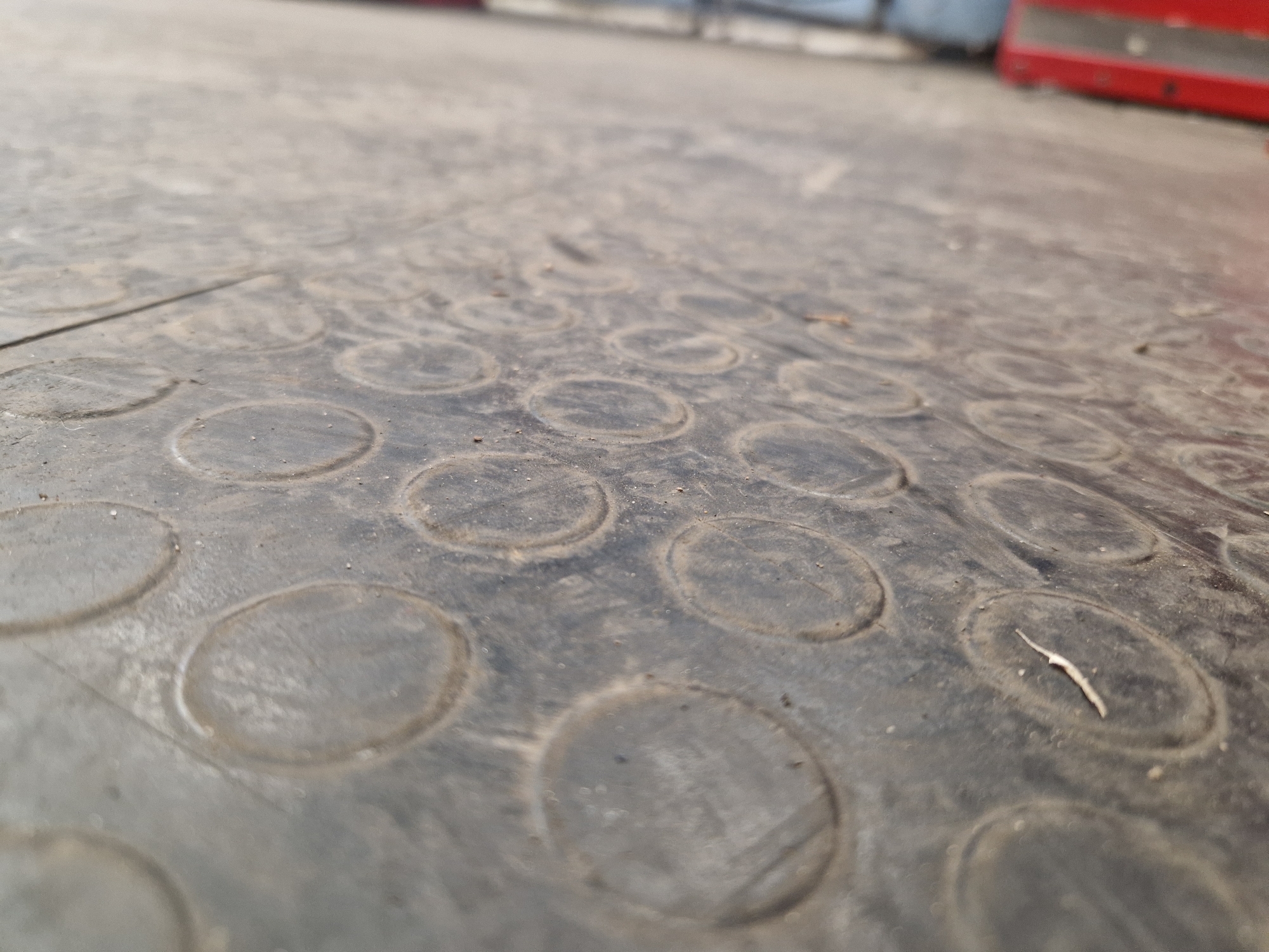 The image displays a close-up, low-angle view of a textured, dark-colored rubber mat or flooring. The surface is predominantly black or very dark grey, covered with numerous small specks of dirt and dust, indicating some wear and use.
The most prominent feature of the mat is its pattern of raised, circular discs, arranged in a grid-like fashion across the visible area. These circles appear to provide anti-slip properties. The texture within each circle seems slightly smoother than the surrounding material, which also shows some grime accumulation. A small, light-colored twig or piece of debris is visible on the mat towards the right side of the frame.
The perspective is very shallow, with the foreground in sharp focus, and the background increasingly blurred. This blur makes it difficult to discern specific objects in the distance. However, hints of shapes and colors suggest an indoor or sheltered outdoor setting, possibly an industrial or utility area. In the upper right quadrant of the background, a distinct blurred red object is visible, which could be part of equipment, a cart, or a container. Other muted, indistinct shapes and tones are present in the blurred mid-ground and background.
No people are visible, nor is any specific activity taking place. The scene is static, focusing on the material and texture of the flooring. There is no visible text. The lighting appears uniform, suggesting a well-lit environment, possibly daytime.