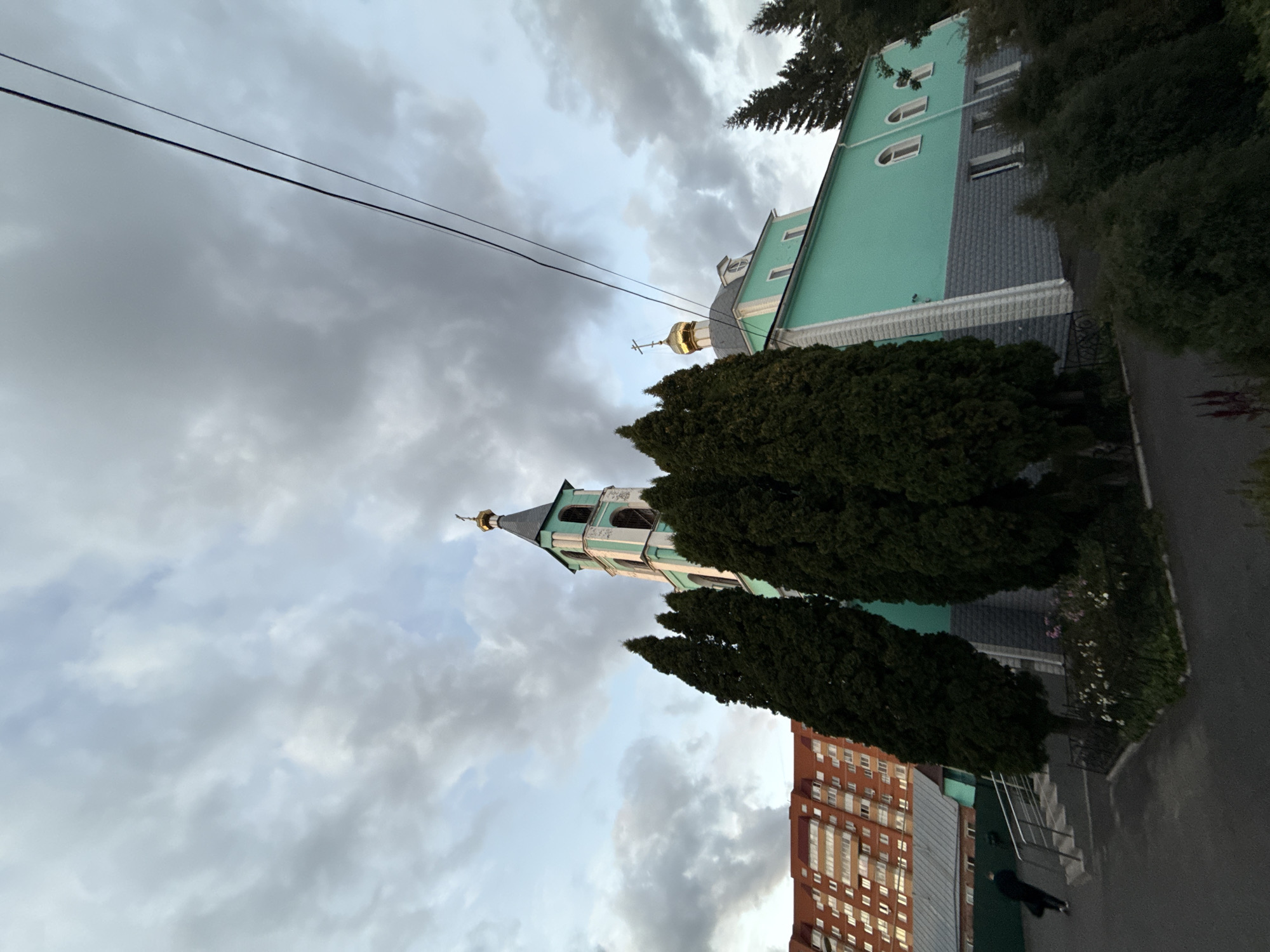 The image shows a turquoise-colored building with a green and gold steeple, characteristic of a Russian Orthodox church, situated in Zheleznogorsk, Russia. The building has arched windows and is partially obscured by tall, dark green coniferous trees. To the left of the church, a multi-story brick apartment building with many windows is visible. The sky above is filled with dramatic, gray clouds, suggesting overcast weather. A person in a dark coat is walking on a paved path leading towards stairs that go up to the church. Power lines are stretched across the upper portion of the frame. The foreground shows a paved area and some greenery, including bushes and flowering plants.