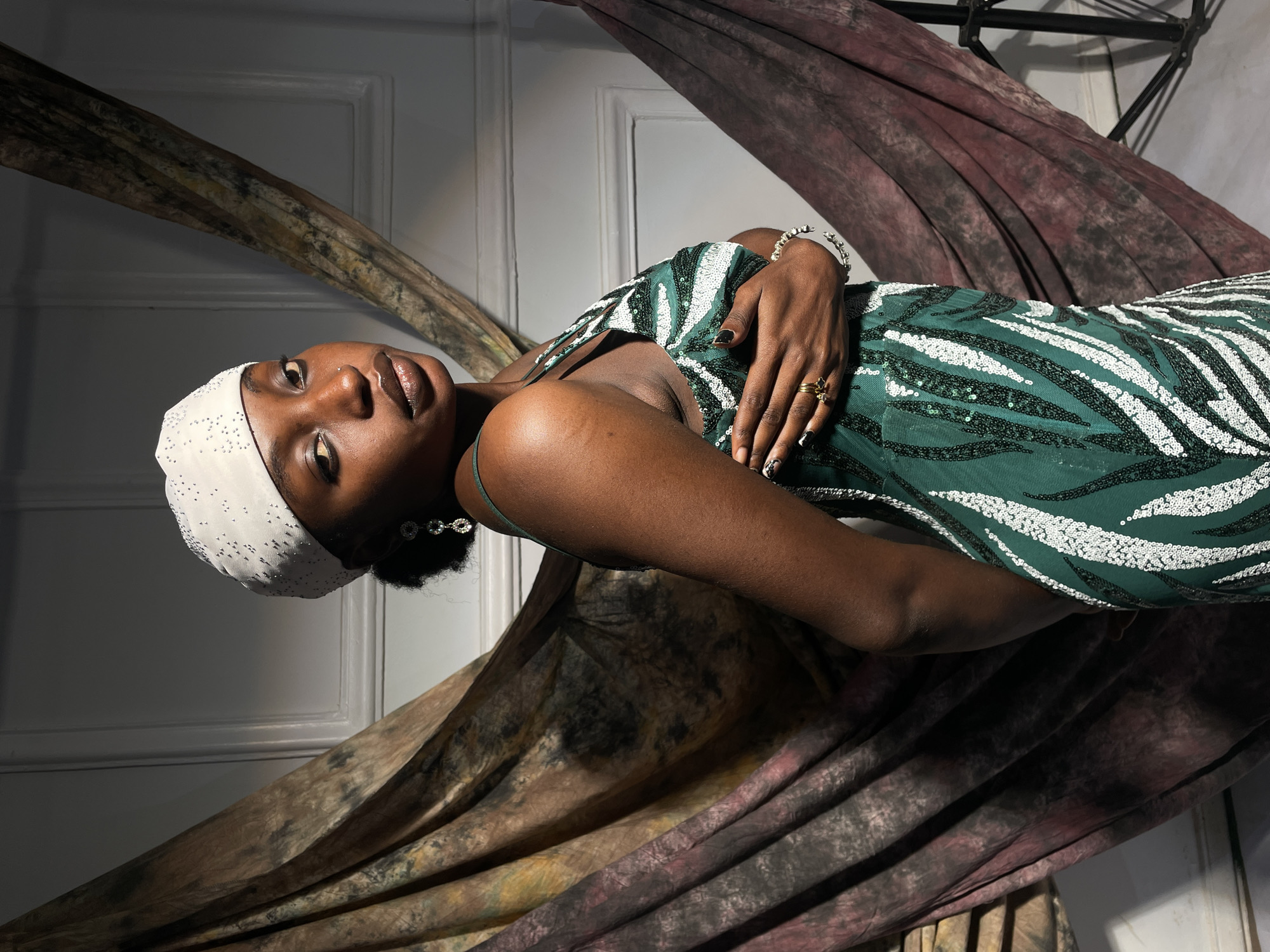 A woman with dark skin is reclining in a posed position against a backdrop of draped fabric, one piece of which is brown and the other a plum color. She is wearing a green and white sequined dress with thin straps, a white cap or head covering with bead-like embellishments, a silver bracelet, and a gold ring. She also has on gold eyeshadow. Her hands are crossed over her body at the center of the frame. The background appears to be an interior setting with a white door visible. The lighting casts shadows, which suggests a controlled, possibly studio-lit environment. The location information is not discernable from the image.