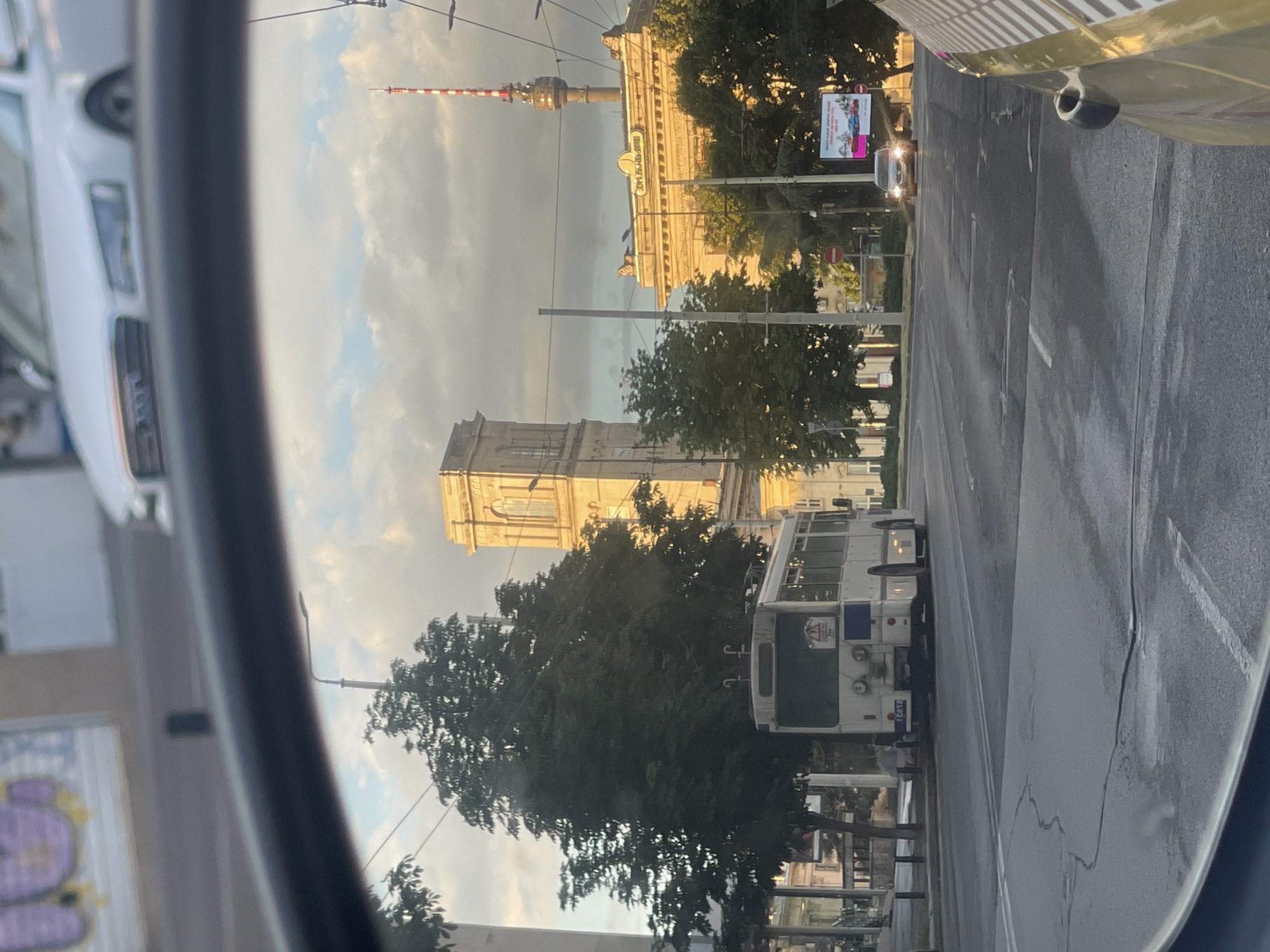 The image, captured from a vehicle's side mirror, depicts a street scene in Ruse, Bulgaria. In the foreground, the dark rim of the mirror is visible, reflecting a cloudy sky. The middle ground is dominated by a white trolleybus on the left, facing away from the viewer, and several large trees. Beyond the trolleybus and trees, a tall, light-colored building with arched windows and a tower is visible. To the right of this structure, another building with a prominent sign and a taller, red-and-white striped radio tower rising behind it. The street is paved and appears wet in places. A few cars are present on the road, including one approaching the viewer and another in the distance. A billboard is visible on the right side of the street. The overall lighting suggests it is either early morning or late afternoon, with the sun casting warm light on the buildings.