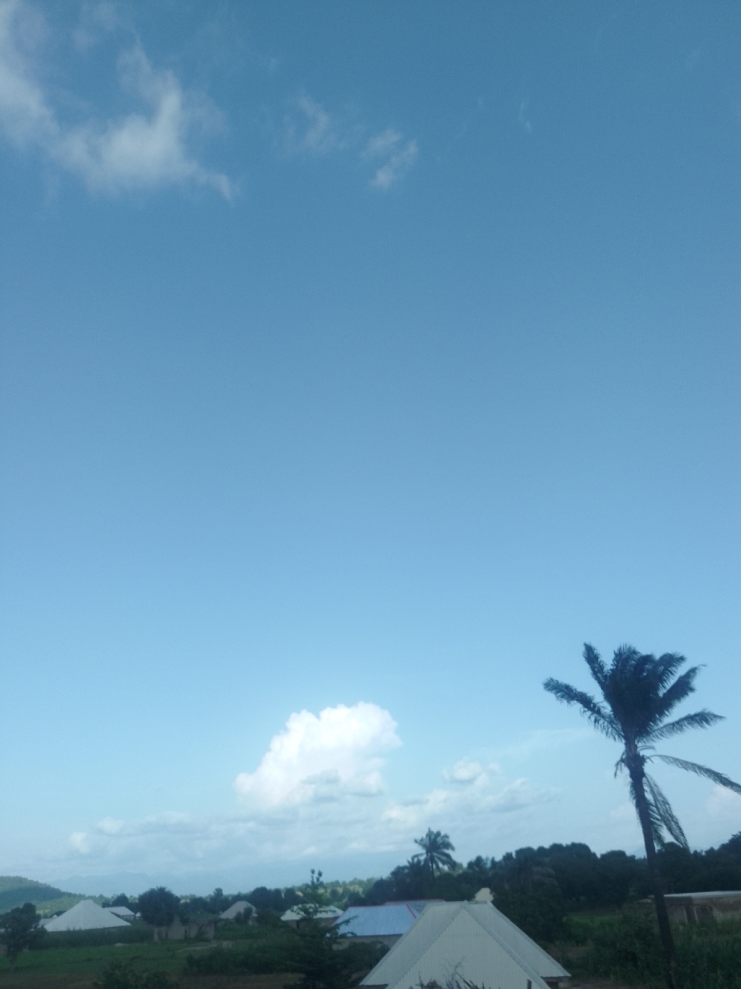 The image shows a bright, clear sky over a rural landscape in Serti, Nigeria. The sky is a vibrant blue, with a few scattered white clouds. In the foreground, there is a tall palm tree with its fronds silhouetted against the sky. Below the palm tree and extending to the left, are several buildings with peaked roofs, some appearing to be white tents or shelters. The surrounding area is covered in green vegetation, with a line of trees in the background and rolling hills further in the distance. The scene appears peaceful and serene. The lighting suggests it is daytime, likely mid-morning or afternoon, given the brightness of the sky. There is no text visible in the image.