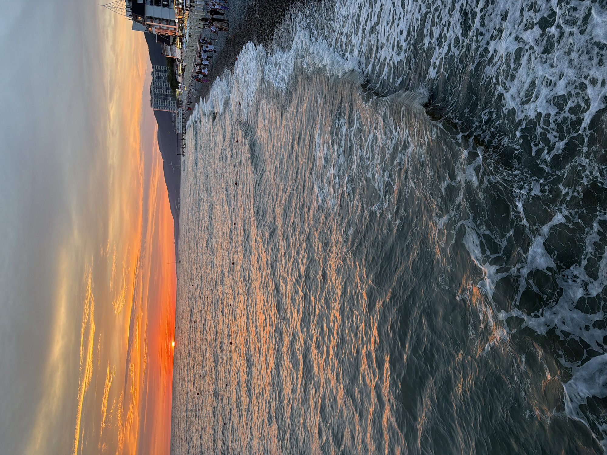This media file depicts a sunset scene at a beach in Sochi, Russia. The image is oriented vertically. In the foreground, waves crash onto a pebble beach. The ocean water is a deep blue-gray, with white foam from the waves. Small, dark objects, possibly buoys or markers, are visible in the water.
The beach itself is lined with people, some sitting on chairs, others standing or walking along the shoreline. Behind the beach, there are several buildings, including apartment complexes and what appears to be a hotel or resort. A dark mountain range serves as a backdrop to the buildings.
The sky is filled with dramatic clouds, illuminated by the setting sun. The sunset casts a warm, orange glow across the clouds and reflects on the surface of the ocean, creating streaks of golden light. The time of day is clearly sunset, indicated by the low angle of the sun and the vibrant colors in the sky. The weather appears to be clear, with the sunset dominating the sky. The presence of buildings and a developed shoreline suggests an urban or resort area. The overall mood evoked is one of a peaceful and beautiful end to the day, with people enjoying the seaside atmosphere. No visible text is present in the image.