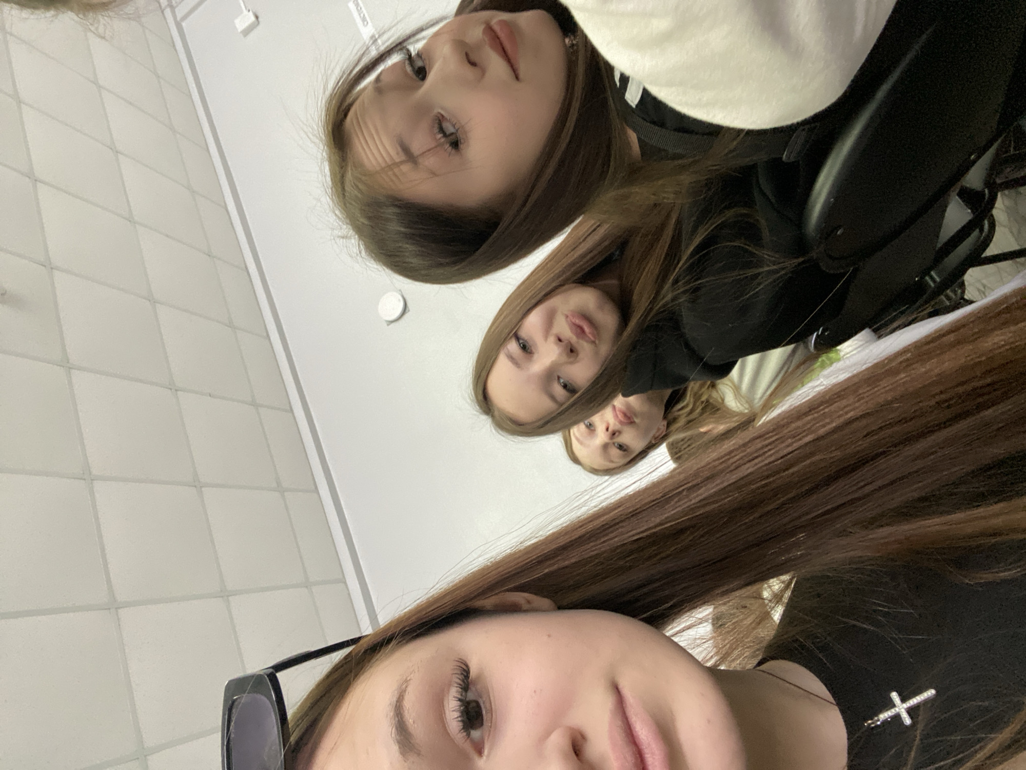 Three young females are posing for a selfie. The background includes a wall with square tiles. The location appears to be indoors. The lower foreground contains the top portion of one female's face, including sunglasses and a silver cross necklace.