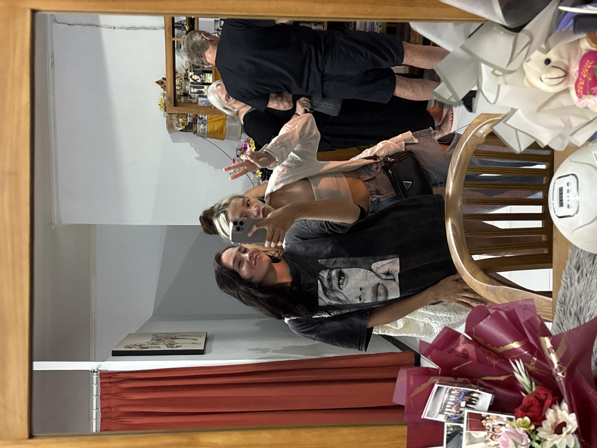 The image shows a group of people in an indoor setting, likely a home in Karangasem, Indonesia. The photo appears to be a selfie taken in a mirror. In the foreground, a young woman with dark hair and bangs is taking the picture. She is wearing a dark t-shirt with a black and white graphic of a woman's face and denim shorts. She is making a peace sign with her right hand.
Behind her, another young woman with blonde streaks in her hair is also visible. She is wearing a white shirt over a light-colored bra and denim shorts. She is holding up her phone and appears to be taking a photo as well.
Further in the background, an older couple is seen. The man is wearing a black t-shirt and shorts, and the woman has short grey hair and is wearing a black dress. They are close together, suggesting an affectionate moment.
The setting includes shelves with various items, possibly decorative or religious objects, and framed photographs. There's a wooden chair and a bouquet of flowers with several small photographs tucked into it. The overall mood seems relaxed and celebratory. The lighting suggests it is daytime or early evening. The presence of a bouquet with photos hints at a possible celebration or special occasion.