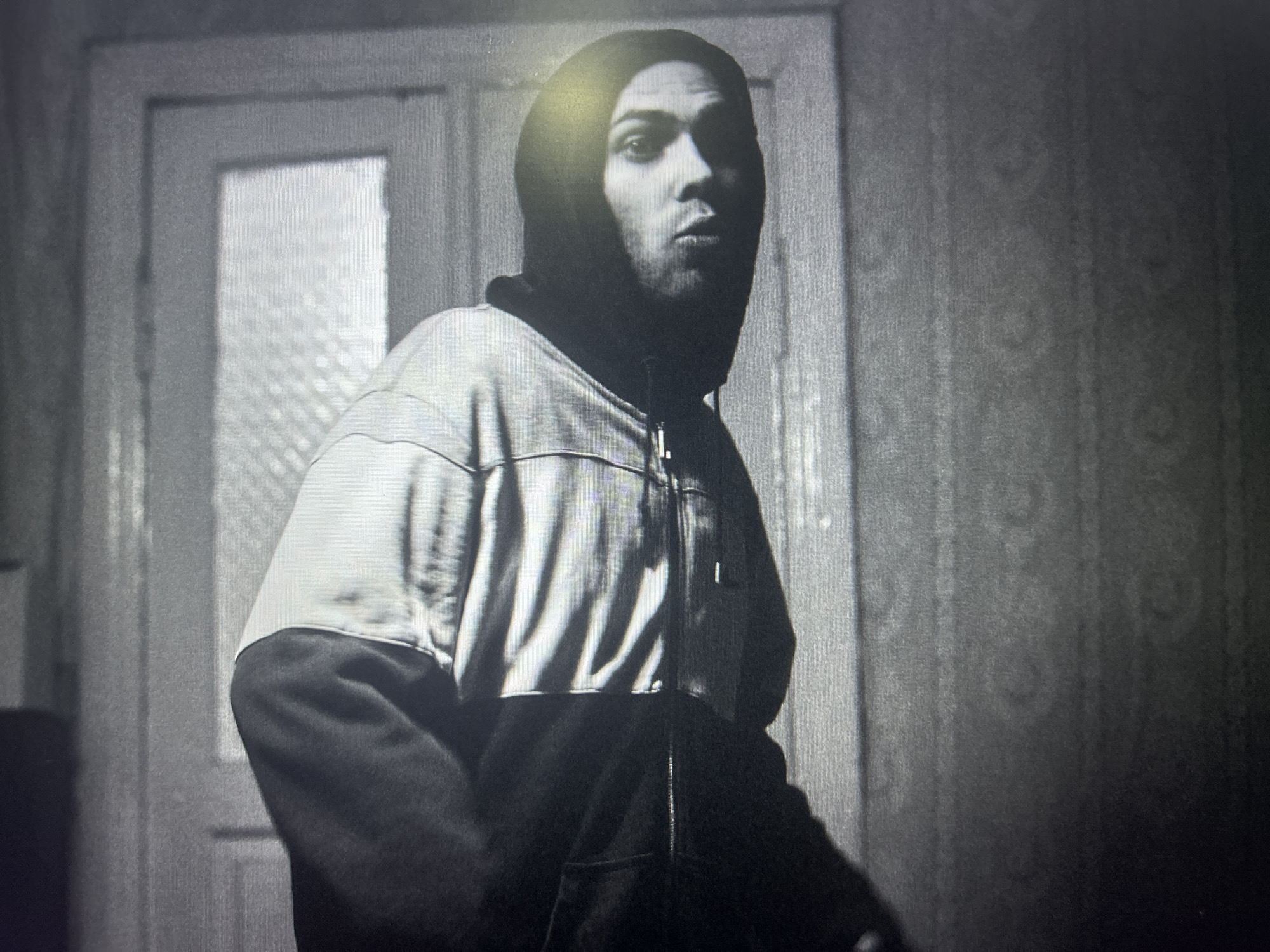 The image depicts a person wearing a hooded sweatshirt, standing indoors. The person is facing the camera with a serious expression. The background includes a door with a frosted glass panel and textured wallpaper. The location is Unknown, Unknown.