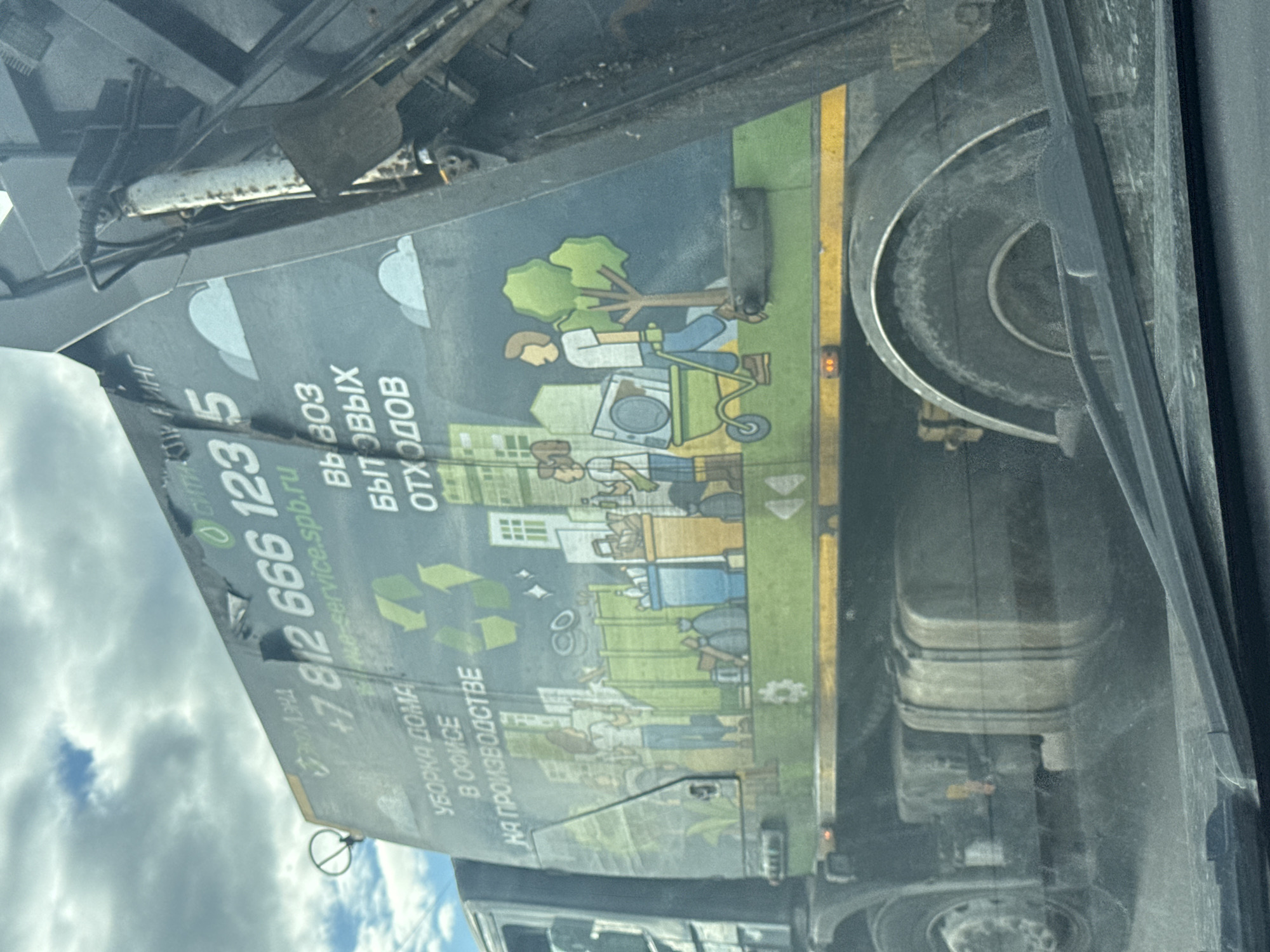 This media file shows the side of a dark-colored truck, likely a garbage truck, with a colorful advertisement graphic printed on it. The graphic depicts stylized people engaged in activities related to waste management and household services. In the upper portion of the graphic, a person is shown tending to a tree next to a washing machine, suggesting a focus on environmental responsibility. Below this, a cityscape with various buildings is illustrated, with people interacting with trash bins and recycling symbols, emphasizing waste collection and recycling. A prominent phone number, "+7 812 666 1235", and a website, "e-service.spb.ru", are clearly visible, along with text in Russian that translates to "Garbage removal" and "Cleaning of homes, offices, and industrial sites". The scene appears to be set during the daytime, with a cloudy sky visible in the background through a reflection on the truck's surface. The overall impression is one of a service company advertising its waste management and cleaning solutions. The image is slightly tilted, as if taken from a moving vehicle.