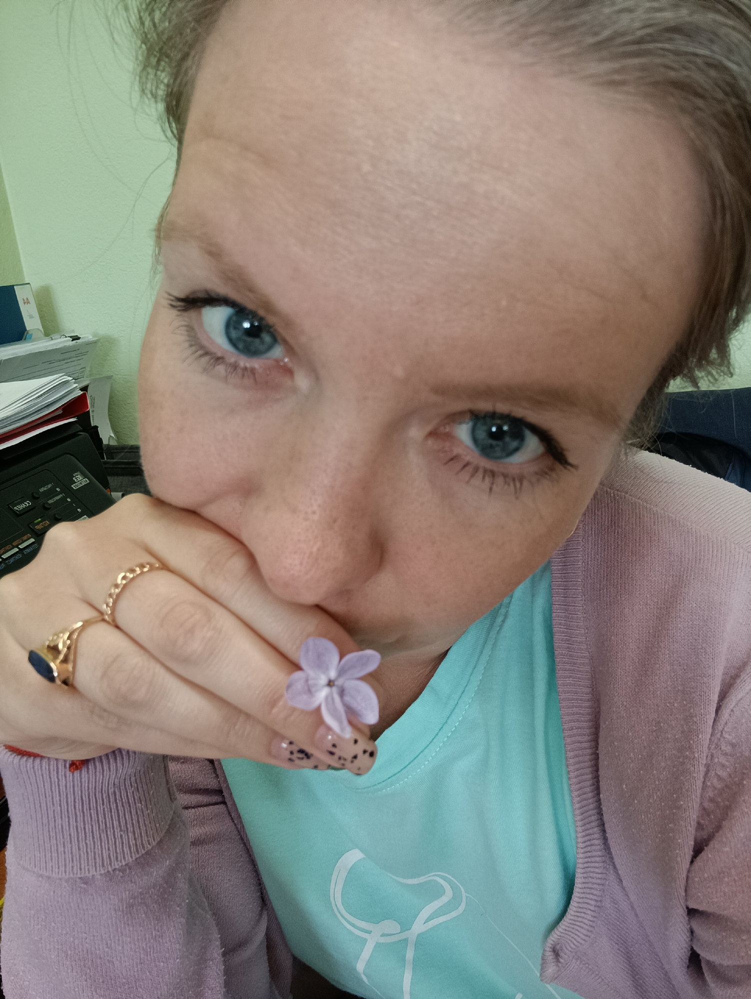 A close-up selfie shows a woman with fair skin and blue eyes. She's holding a small purple flower close to her mouth with her left hand. She is wearing two gold rings on her left hand. Her fingernails are painted with a clear base coat and black dots. She is wearing a light blue t-shirt and a light pink cardigan. The background consists of office clutter, including a phone and paperwork. The location is identified as Chaikovsky, Russia.