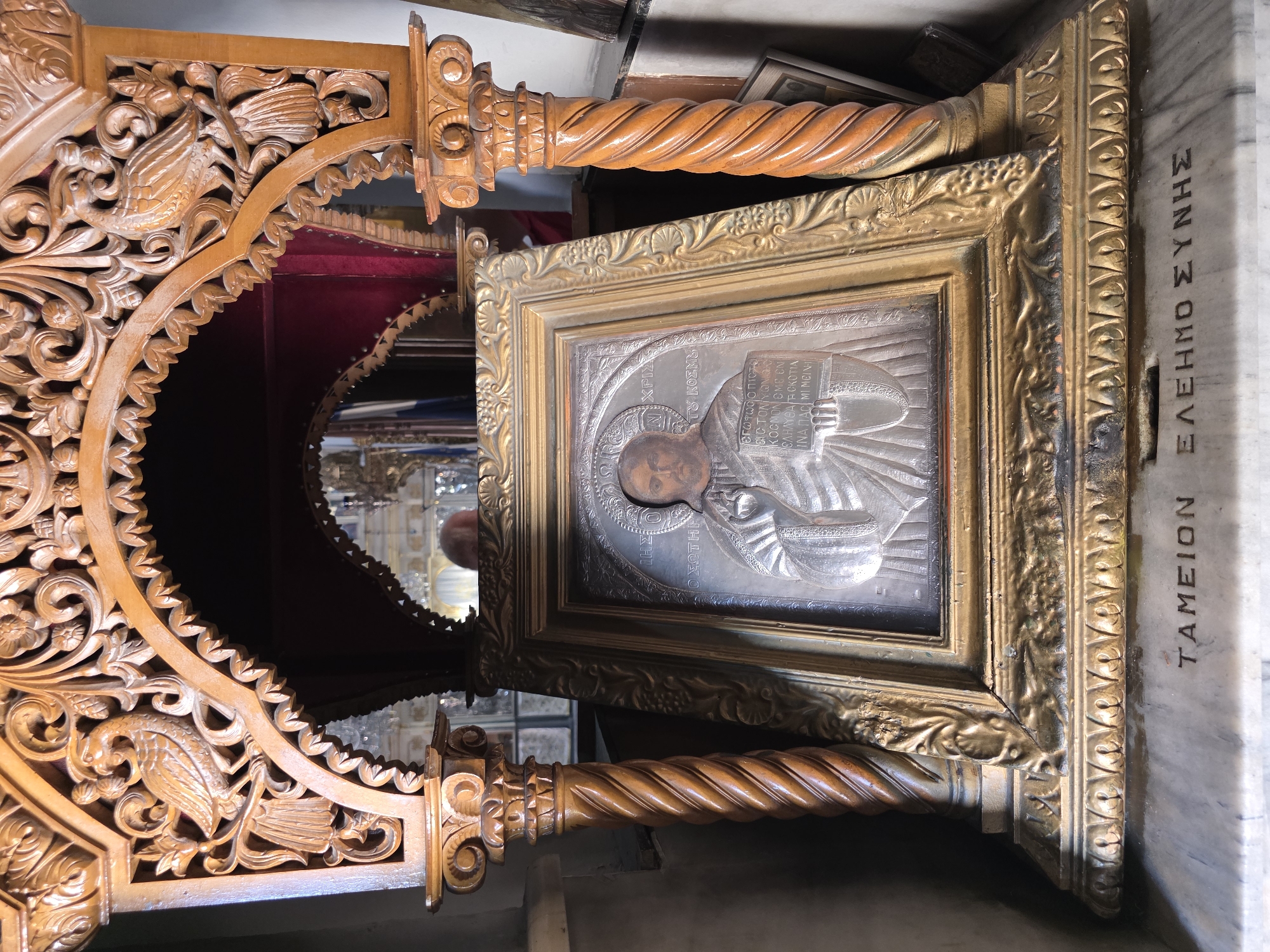 The image depicts an Orthodox Christian icon of Jesus Christ, framed in ornate gold, positioned within a wooden, carved shrine. The shrine features intricate carvings of birds and floral motifs, with twisted columns on either side. The icon itself is made of repoussé metal, likely silver, depicting Christ with a halo, holding a book and making a gesture of blessing. In the background, a red velvet curtain is visible, and through an archway, a reflection shows a portion of the church interior, including a chandelier and the head of a person. Below the icon, on a marble surface, is an inscription in Greek letters that reads "TAMEION ELEHMO SYNHΣ". This inscription translates to "Charitable Fund" or "Mercy Fund". This scene is located in the Saint Titus Cathedral in Chania, Greece.