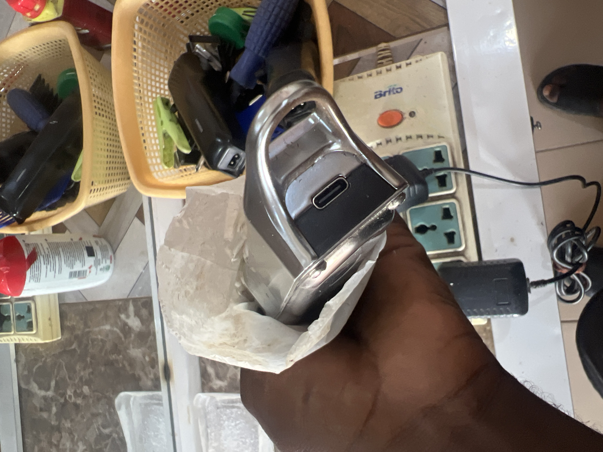 In Jalingo, Nigeria, a close-up shot reveals a hand holding a device wrapped in a white paper bag. The device, appearing to be a small electric clipper or trimmer, has a metallic handle and a USB-C charging port. In the background, a power strip with multiple outlets is visible, along with two yellow plastic baskets filled with various grooming tools, including clippers, brushes, and hair clips. A bottle with a red cap and white label sits on a tiled surface. The overall environment suggests a barber shop or a personal grooming space.