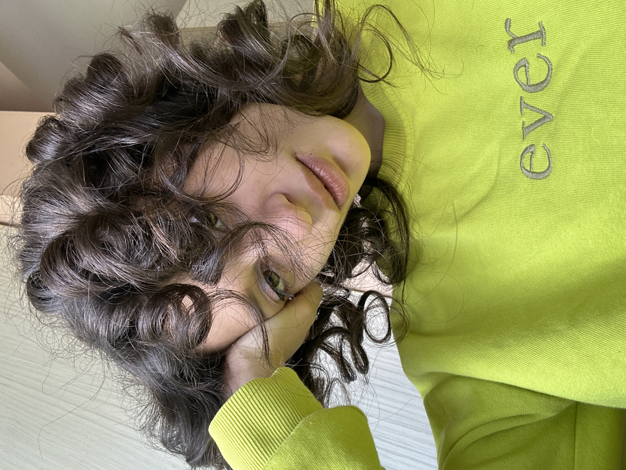 The image is a selfie of a young woman with dark, curly hair and light-colored eyes. She is wearing a bright yellow-green sweatshirt with the word "ever" embroidered in grey on the front. Her head is resting on her hand, and she is looking directly at the camera. The background appears to be a wall with a subtle texture. There is no identifiable location in Sofia, Bulgaria visible in the image.