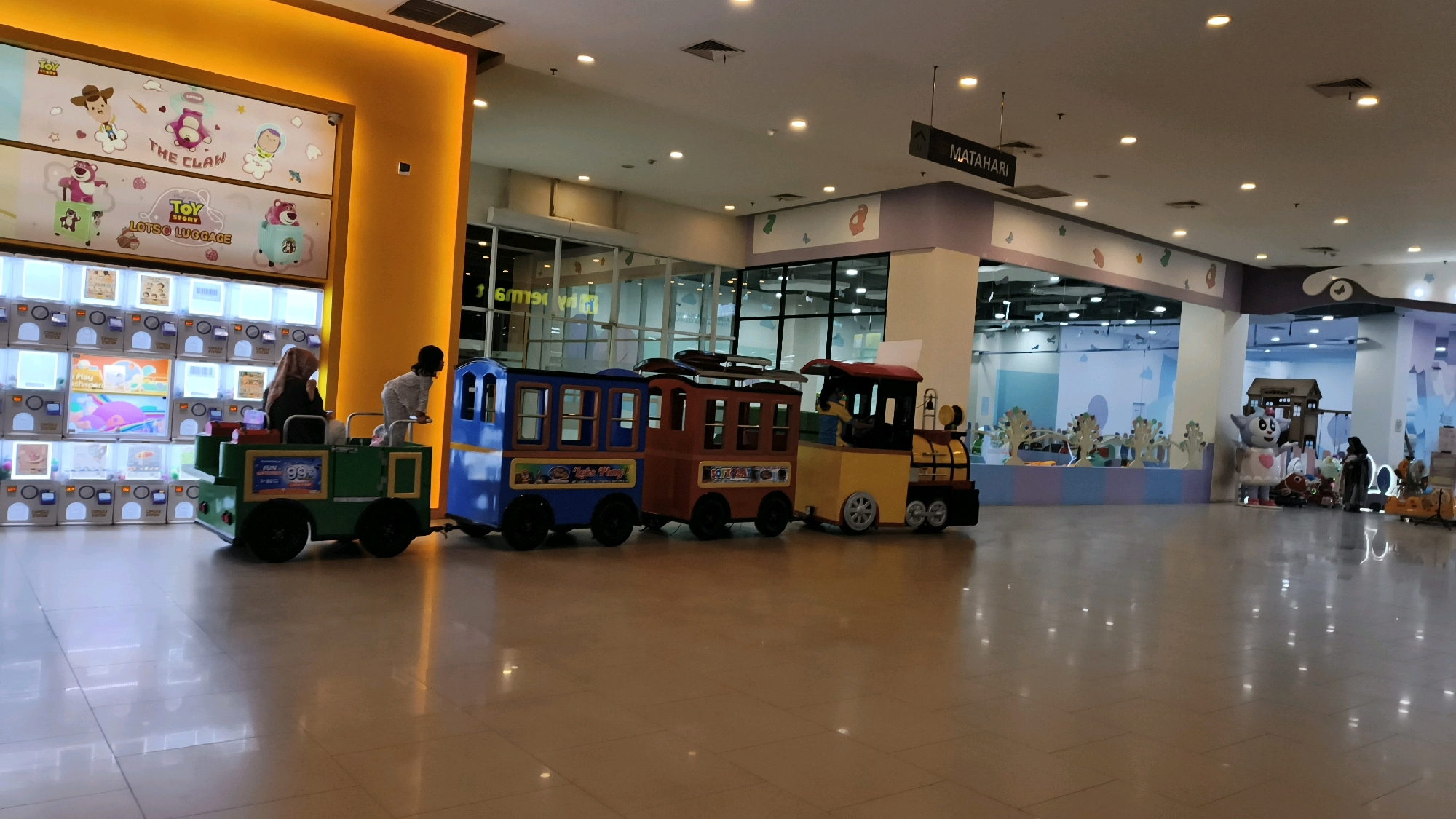 This image captures an indoor amusement area, likely a children's play zone within a shopping mall in Baturaja, Indonesia. In the foreground, a colorful toy train with three carriages is parked on the polished floor. The train is green, blue, and brown with a yellow engine. Two people are partially visible, one seated in the green carriage and another standing in the blue carriage. To the left, a wall features a display of numerous vending machines, many illuminated, with graphics related to "Toy Story" and "The Claw" on the upper section. The background reveals a brightly lit play area with decorative elements, including cartoon characters, trees, and a small building structure. A sign labeled "MATAHARI" hangs from the ceiling, indicating a department store. The environment is clean and well-maintained, with reflections on the floor suggesting a smooth, possibly tiled surface.