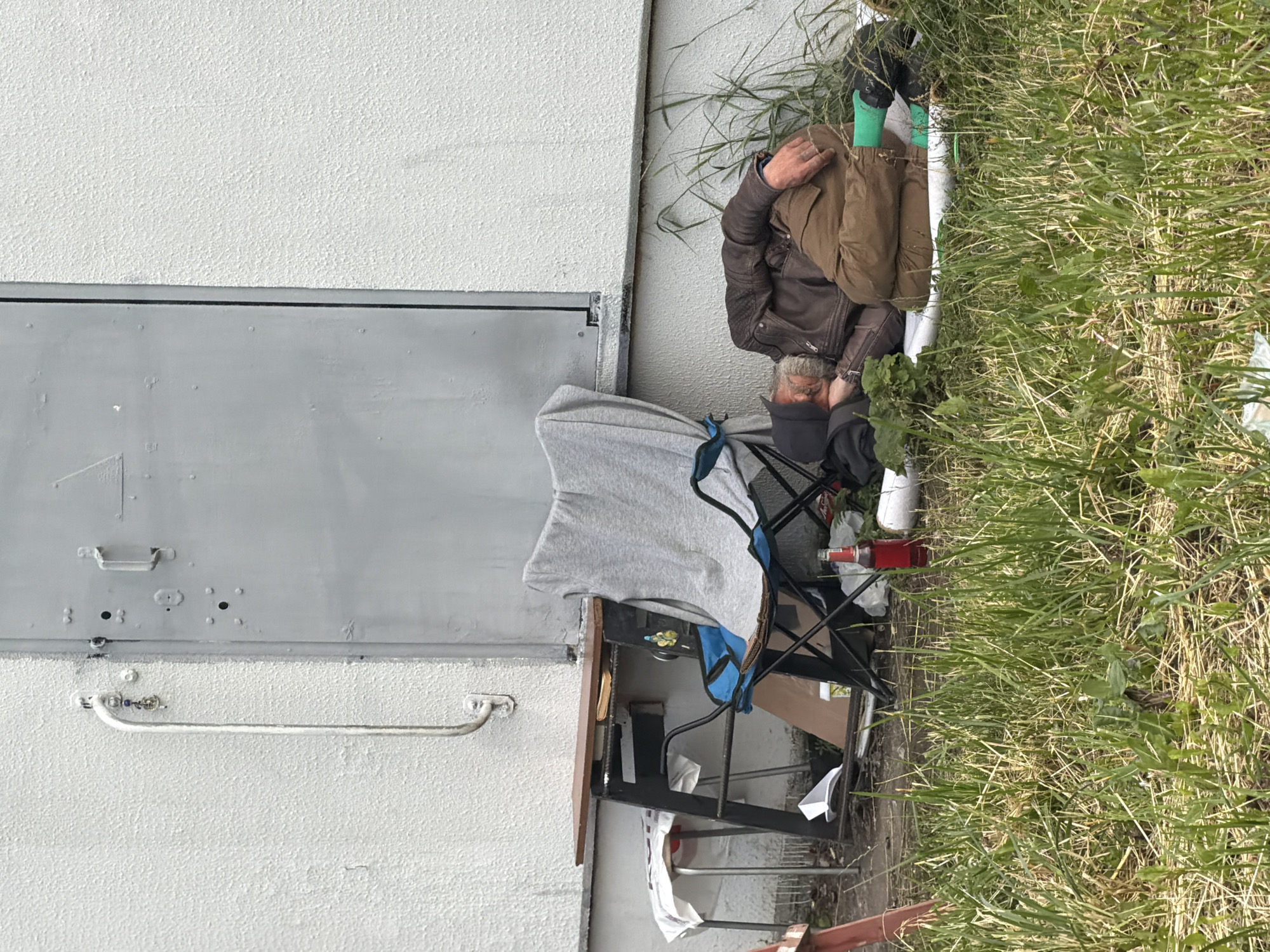 An adult male is sleeping outdoors, lying on his right side on a white sheet in the grass. He is wearing a dark-colored baseball cap, a brown leather jacket, brown pants, and green socks. He has gray hair and a beard. Next to him is a folding camp chair covered with a grey blanket. Also visible are a red bottle, some green foliage, and a portion of a white building with a large gray door. A small wooden table with a metal frame stands to the left of the chair. A white stool covered with a cloth is near the table. The scene is illuminated by natural daylight.