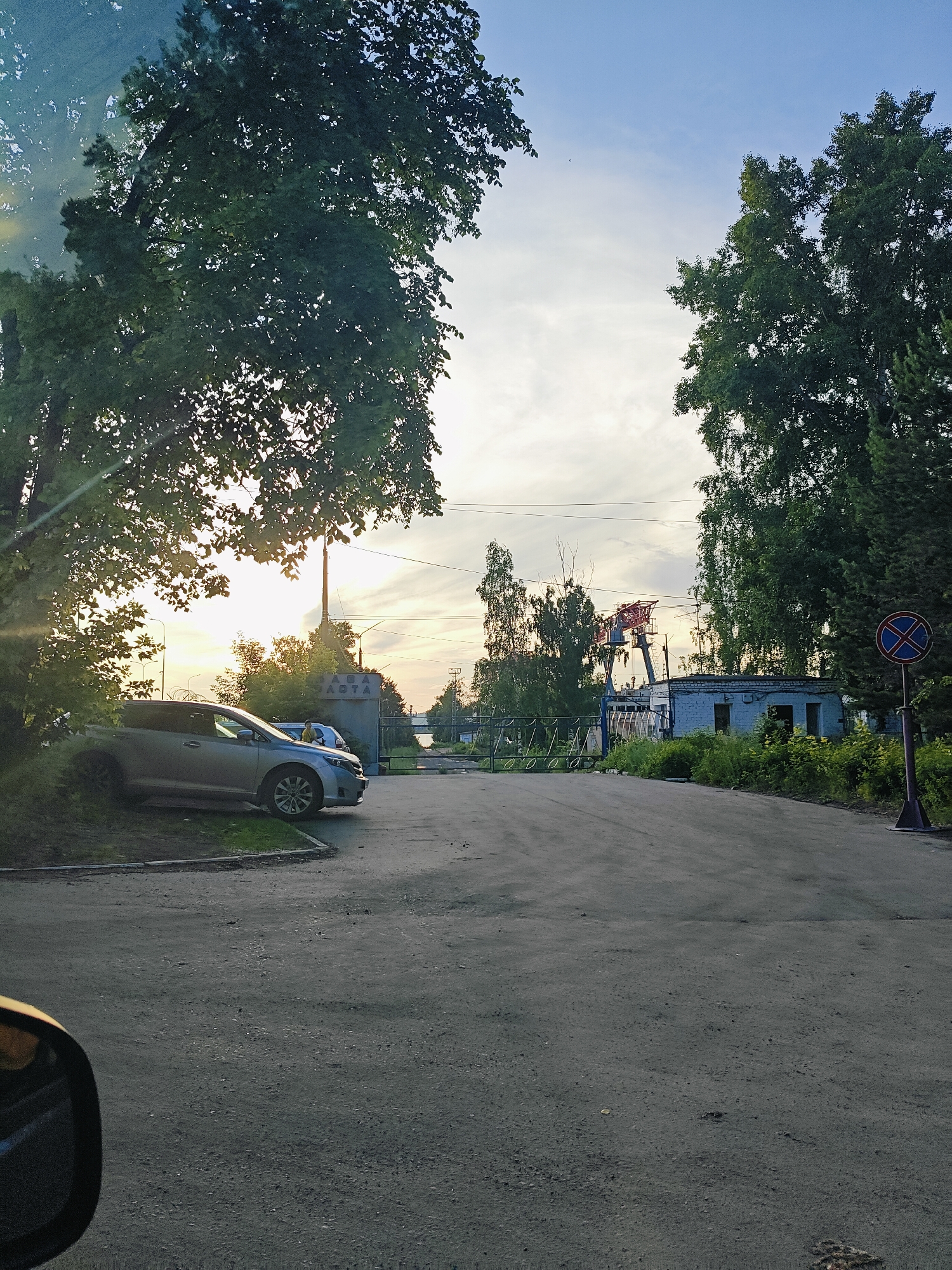 The image depicts an outdoor scene in Chaikovsky, Russia, likely in the late afternoon or early evening, as indicated by the warm, low-angled sunlight and the long shadows. The setting appears to be an industrial or semi-industrial area, with a paved road leading into the frame.
In the foreground, on the left, a silver car is parked partially on the grass verge. The car is a modern sedan. To the right, the road continues into the distance, flanked by overgrown greenery and trees. In the middle ground, there is a closed metal gate, beyond which stands a low, light blue building with a white roof. Behind the building, a tall red and grey industrial crane is visible, suggesting some form of manufacturing or construction activity. A dark blue circular sign with a red diagonal line and a red cross within it is mounted on a pole on the right side of the road, indicating a prohibition of some sort, likely related to parking or stopping.
The sky is partly cloudy, with the sun filtering through the clouds, creating a hazy, warm light. The overall mood is serene, with a sense of quietness pervading the scene. The presence of industrial elements contrasts with the natural setting of trees and greenery. Visible text on a structure behind the gate reads "АВТО".