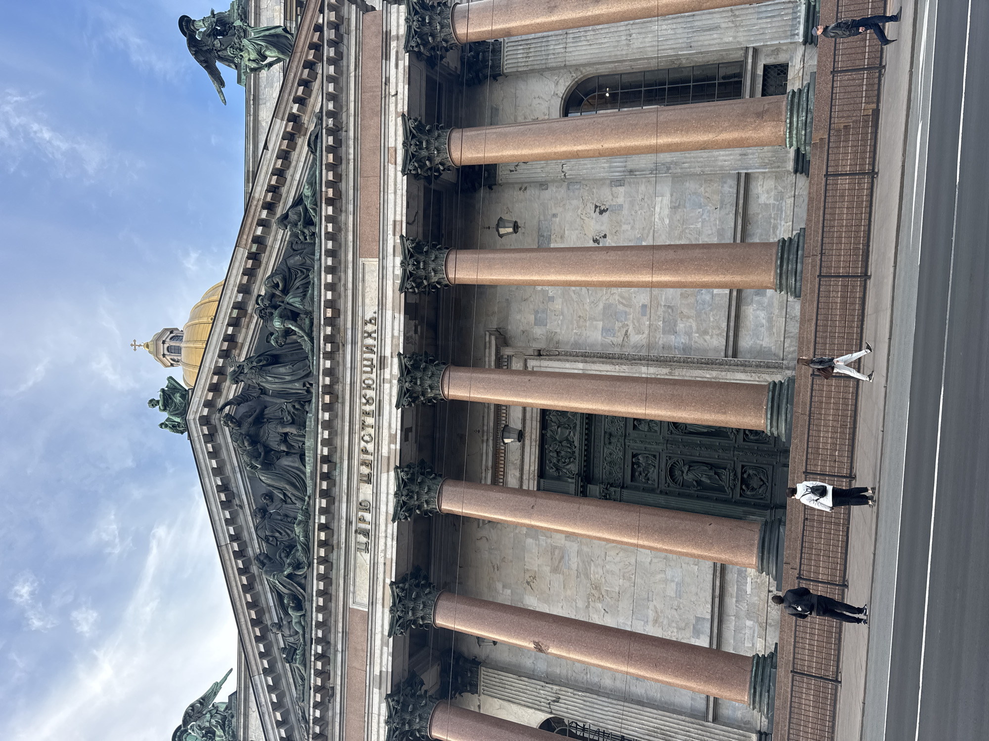 An outdoor, upward-angled shot captures the grand facade of a massive cathedral, identified as St. Isaac's Cathedral in Saint Petersburg, Russia. The building dominates the frame with its imposing architectural features.
The facade is characterized by a colonnade of enormous, reddish-brown granite columns with intricately carved, dark capitals. Above these columns, a frieze bears Cyrillic text, partially visible as "ГЛАГОЛЫ СВѦТЫХЪ" (Words of the Saints), and beyond that, a pediment adorned with numerous dark, weathered sculptures of figures. Higher up, additional statues can be seen along the roofline, against a bright blue sky with scattered white clouds. The building's golden dome, topped with a cross, is visible in the upper left, partially obscured by the foreground architecture. Large, ornate dark green doors are recessed between the columns, leading into the building.
Below the columns, a sidewalk runs parallel to the structure, separated by a dark metal fence. Several people are seen on the sidewalk. On the right, a person in a dark jacket walks towards the foreground, while another in a white jacket and dark pants stands closer to the fence. Further back, two more individuals are visible, one in a dark outfit and another in lighter clothing. The scene suggests a pleasant daytime, with clear weather. The lighting is bright and even, indicating either late morning or early afternoon. The people appear to be casually traversing the area, perhaps as tourists or pedestrians.