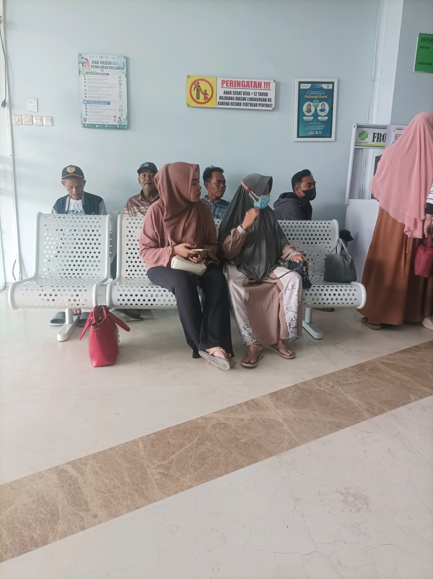 The image shows a waiting area in what appears to be a medical facility, possibly a hospital or clinic, in Sumuragung, Indonesia. Several people are seated on white perforated metal chairs, indicating a waiting period. A red handbag is placed on the floor in front of one of the chairs. There are posters on the wall with text in Indonesian, including what appears to be health information and rules. The flooring is a light-colored marble with a contrasting darker marble stripe. Natural light seems to be coming from the left side of the frame. The overall atmosphere is that of a public waiting space.