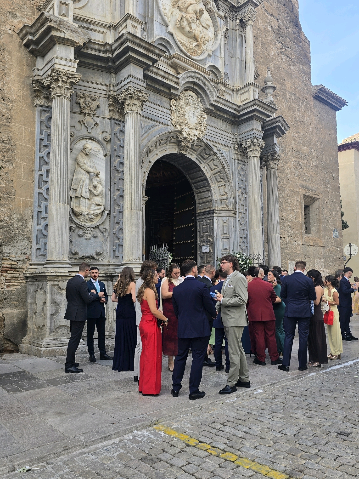The image depicts a gathering of people in formal attire outside a historic building, likely a church or cathedral, in Granada, Spain. The architecture is characterized by ornate stone carvings, columns, and a large arched doorway. The people are dressed in suits and evening gowns, suggesting a special occasion such as a wedding or formal event.
Specifically, the scene appears to be in front of the Alhambra or a similar historic monument in Granada. The stone facade and the architectural style are indicative of Moorish and Renaissance influences common in the city. The cobblestone street and the bright, clear sky suggest a warm, sunny day.
The individuals are standing around, some in small groups engaged in conversation, while others are looking towards the entrance of the building. The overall atmosphere is one of elegance and festivity. There's a floral arrangement near the entrance, adding to the celebratory mood. The composition of the image captures both the grand architecture and the social gathering.