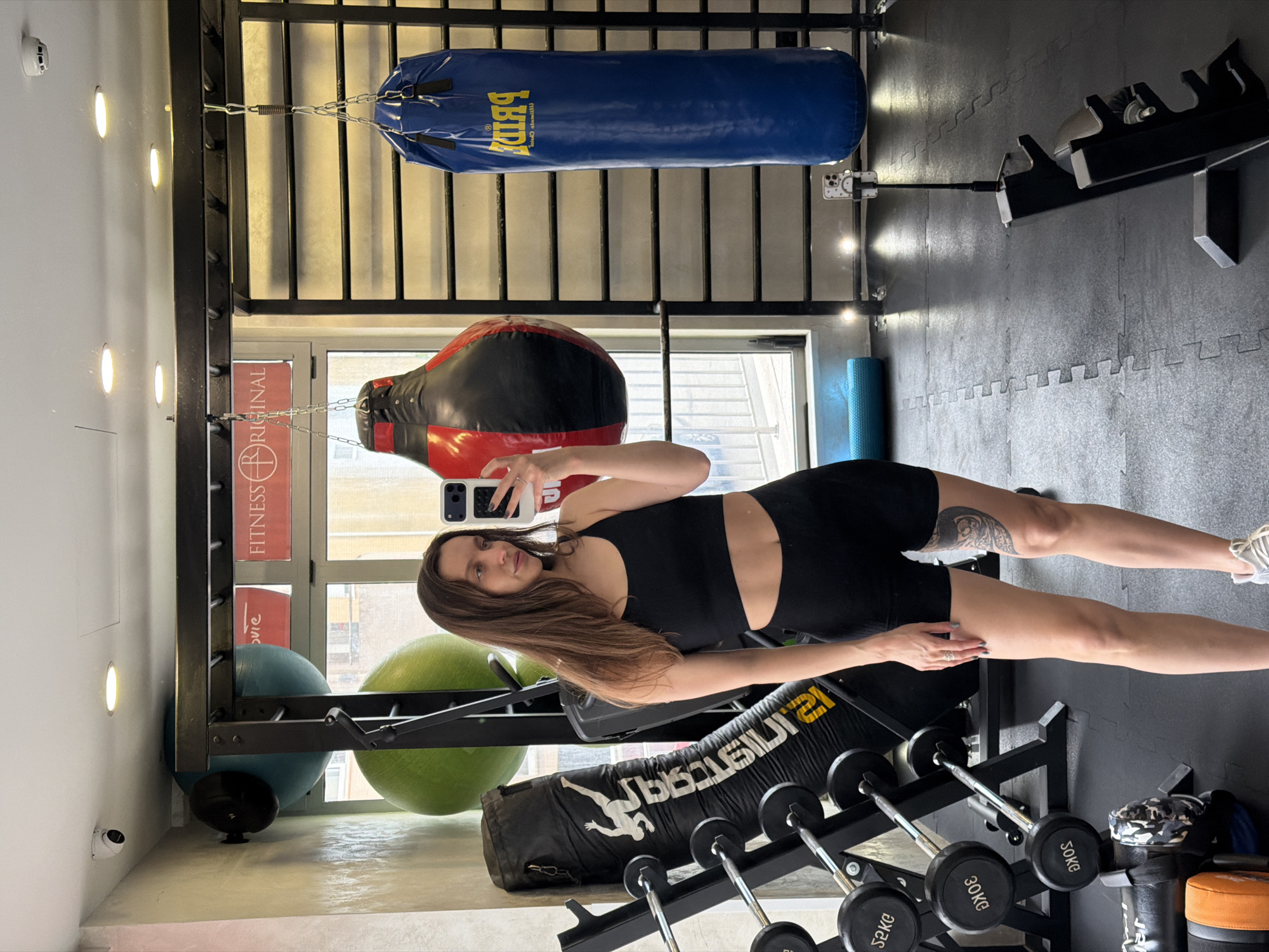 A woman in black sportswear is taking a selfie in a gym. The gym is located in Budva, Montenegro, as inferred from the context (assumed, not explicitly stated in the image). The environment includes various gym equipment such as punching bags, exercise balls, weights, and weight racks. The woman is holding her phone up to take the photo, and her arm is extended to capture the shot.