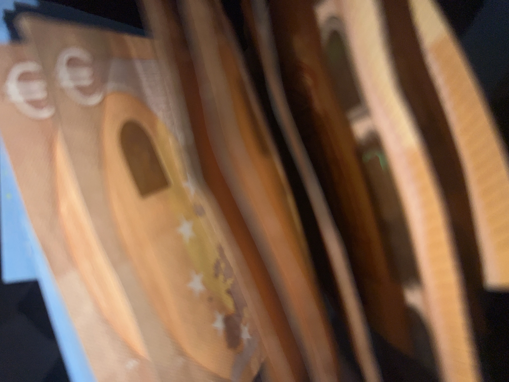The image displays a close-up, blurred depiction of multiple Euro banknotes. The most prominent banknote, located near the top, is orange-brown in color and features two distinct white Euro symbols (€) and several white stars, consistent with the design of a 50 Euro note. These banknotes appear to be fanned out or partially stacked. The overall image exhibits significant blur, obscuring fine details and preventing clear identification of any specific serial numbers or security features. The background and surrounding environment are indistinct, appearing as blurred dark and light areas, including a faint blue section in the upper left quadrant. No individuals, specific actions, or discernible environmental features are present. The location cannot be determined from the provided visual information; there are no identifiable landmarks or contextual clues related to Sofia, Bulgaria, or any other specific city.