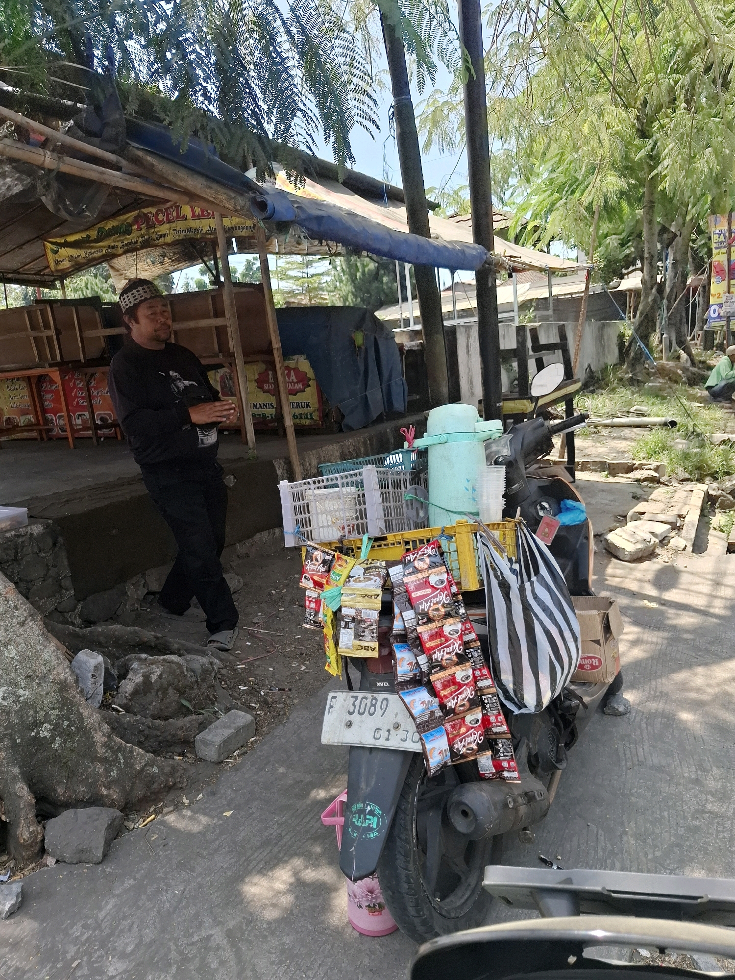 An outdoor scene depicts a man standing beside a scooter loaded with various items, possibly in Sukalarang, Indonesia. The man, wearing a black long-sleeved shirt, black pants, and a patterned skullcap, stands with his legs crossed and hands clasped. He appears to be looking to his left. Behind him, a simple wooden structure with a tarp roof forms a stall, displaying signs with text in Indonesian. The scooter, parked on a paved surface, has a large number of individually packaged items, likely food or drink, attached to its front and sides. These packages are colorful and varied, some featuring text like "Kopi ABC" and "Kopi Gula". A black and white striped bag hangs from the scooter's handlebars. A large tree trunk with exposed roots is visible to the left of the scene, with scattered rocks and debris on the ground. The background shows more greenery and structures, suggesting an area with both natural elements and human settlement. The lighting suggests it is daytime.