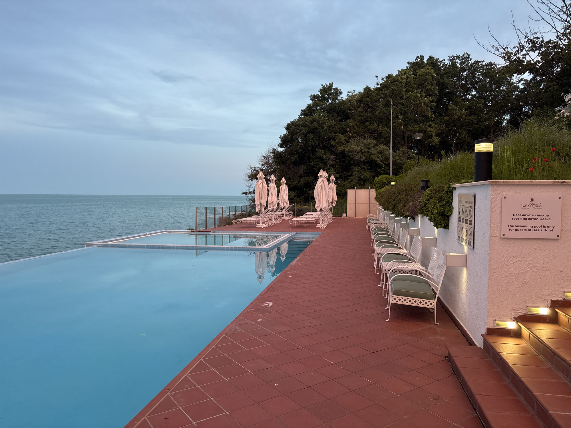 The image shows an outdoor infinity swimming pool with clear blue water, reflecting the cloudy sky. The pool area is paved with red tiles. To the right of the pool, there is a row of white lounge chairs with green cushions, and tables. Further to the right, there is a white building with stairs leading up, illuminated by warm lights. Green bushes and trees are visible in the background. A sign on the building states "The swimming pool is only for guests of Oasis Hotel". The ocean is visible to the left of the pool. The scene appears to be at dusk. This is the outdoor swimming pool area of Oasis Hotel, located on the coast of Varna, Bulgaria.