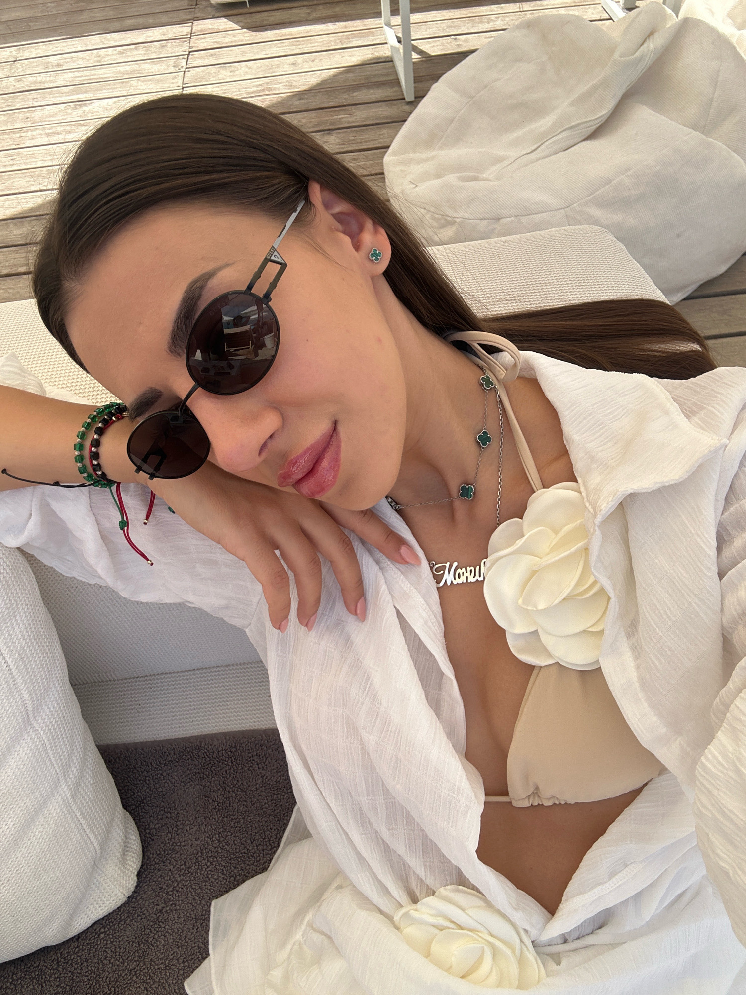 A young woman with dark hair, wearing sunglasses, a white sheer cover-up over a bikini top, and jewelry, rests her head on her arm. She is lying on a sun lounger on a wooden deck. In the background, a large, light-colored beanbag chair is visible. The scene suggests an outdoor leisure setting, possibly a poolside or beach area, consistent with the coastal environment of Pallini Municipal Unit, Greece.