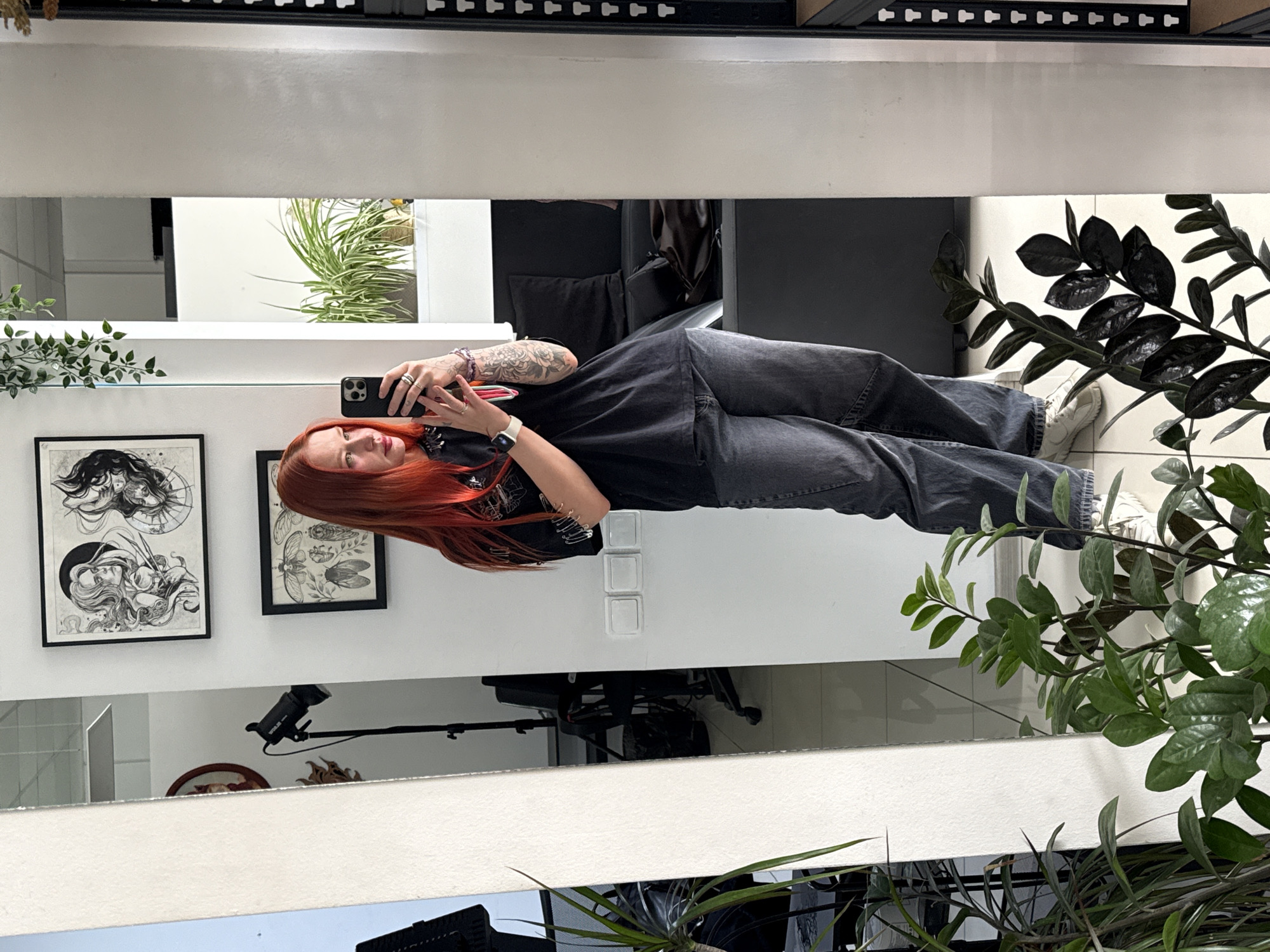 A redhead woman takes a selfie in a mirror. She wears a black t-shirt and dark gray jeans. Her left arm has a tattoo. Behind her are framed artworks on the wall, and potted plants. The reflection in the mirror shows a studio light. Based on the provided images, there's no evidence to conclude this image was taken in Rostov-on-Don, Russia.
