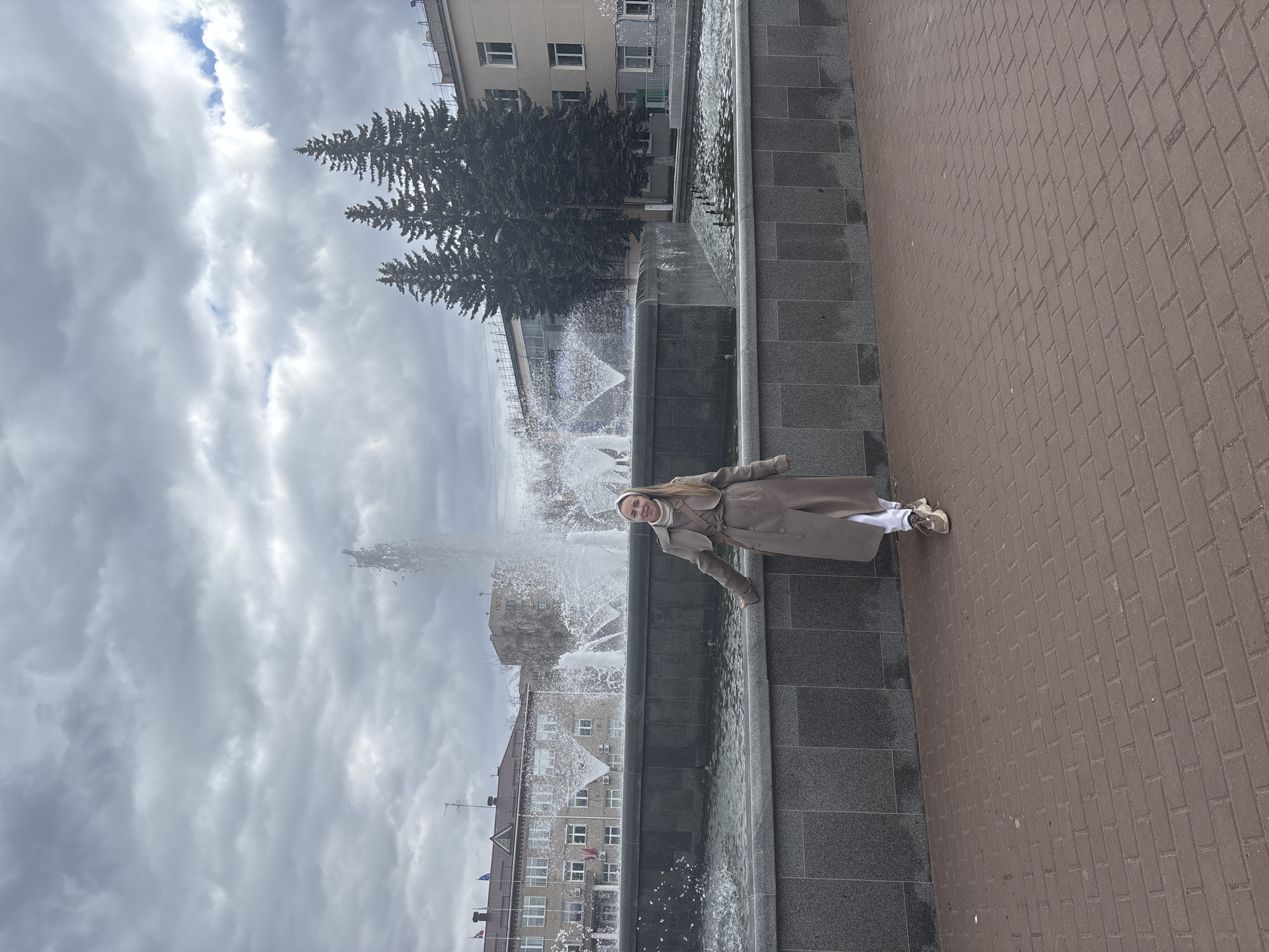 A woman stands next to a large fountain in Chaikovsky, Russia. The fountain is made of dark gray stone and has multiple tiers with water spraying upwards. Buildings are visible in the background, along with tall evergreen trees. The sky is overcast with gray clouds. The woman is wearing a tan coat and white shoes. She is positioned to the right of the fountain. The pavement to the right of the fountain is made of red bricks.
