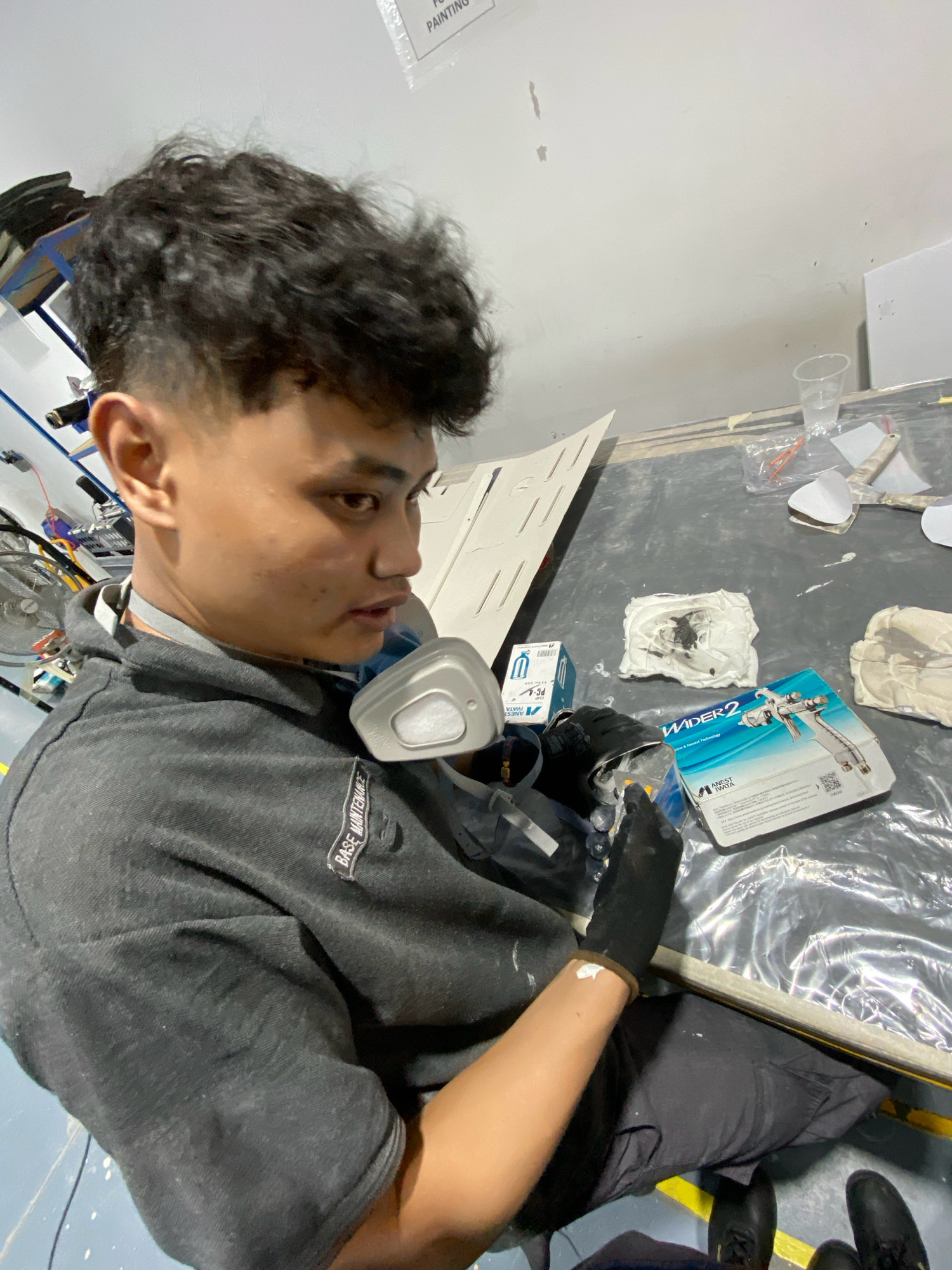 The image shows a young man wearing a grey hoodie and a respirator mask, working on a project. He is wearing black gloves and appears to be applying some sort of material. On the table in front of him are various items, including a box for an Anest Iwata WIDER2 spray gun, a small container of white material, and some other tools. The background suggests an indoor workspace, possibly a workshop or industrial area, with shelves and equipment visible. The overall scene depicts a hands-on activity, likely related to painting or repair. The question about the location in Batam, Indonesia is not answerable from the image itself.