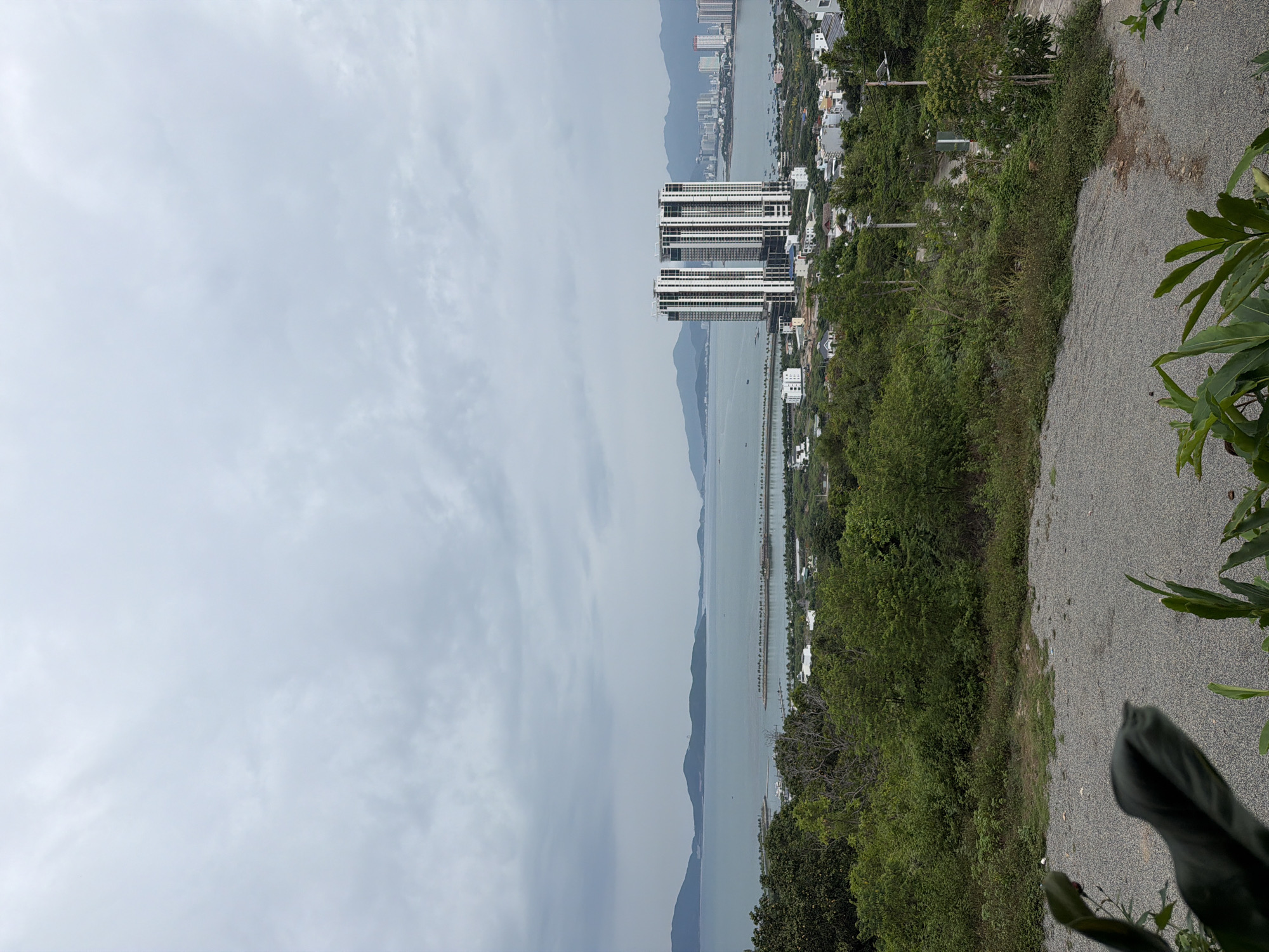 The image shows a view from an elevated position in Bac Nha Trang Ward, Vietnam. The sky is overcast with pale gray clouds. In the distance, a body of water extends with mountainous terrain visible on the opposite shore. A large white high-rise building is centrally located, positioned along the waterfront. Closer to the foreground, there is dense green vegetation including trees and shrubs. The edge of a gravel path or road is visible on the right side of the image.
